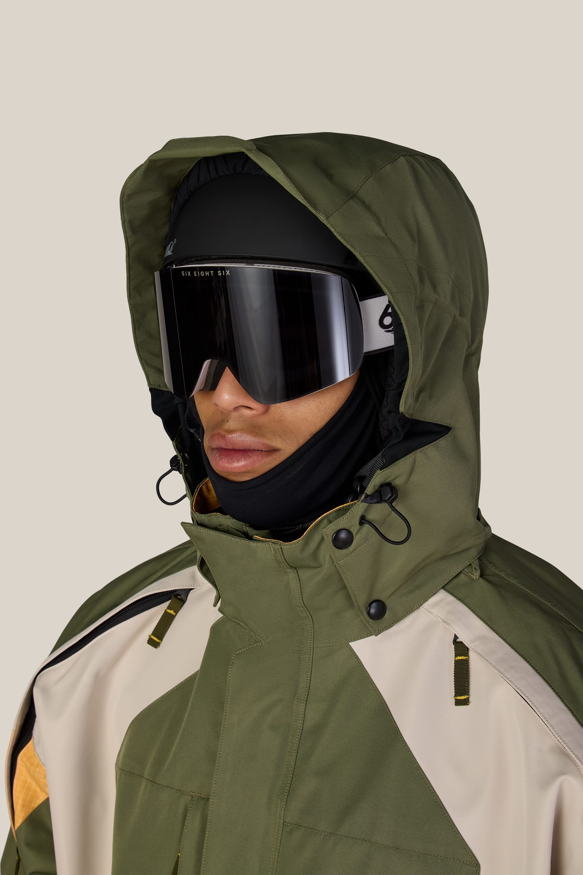 A person wearing the 686 Men’s 2003 Atom Jacket in olive green with a hood, paired with black ski goggles and a black balaclava, facing slightly to the left against a plain light background.