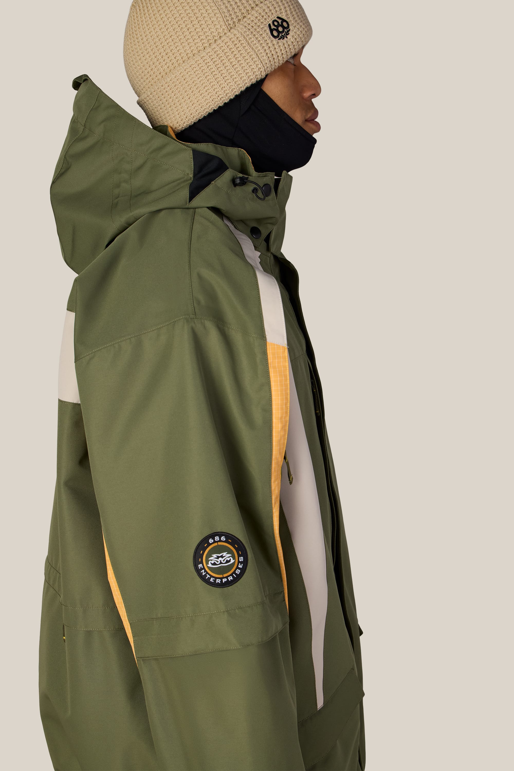 A person is shown in profile against a plain light background wearing the 686 Men’s 2003 Atom Jacket—a green waterproof jacket with yellow and white accents from 686—paired with a beige knit beanie and a black neck covering.