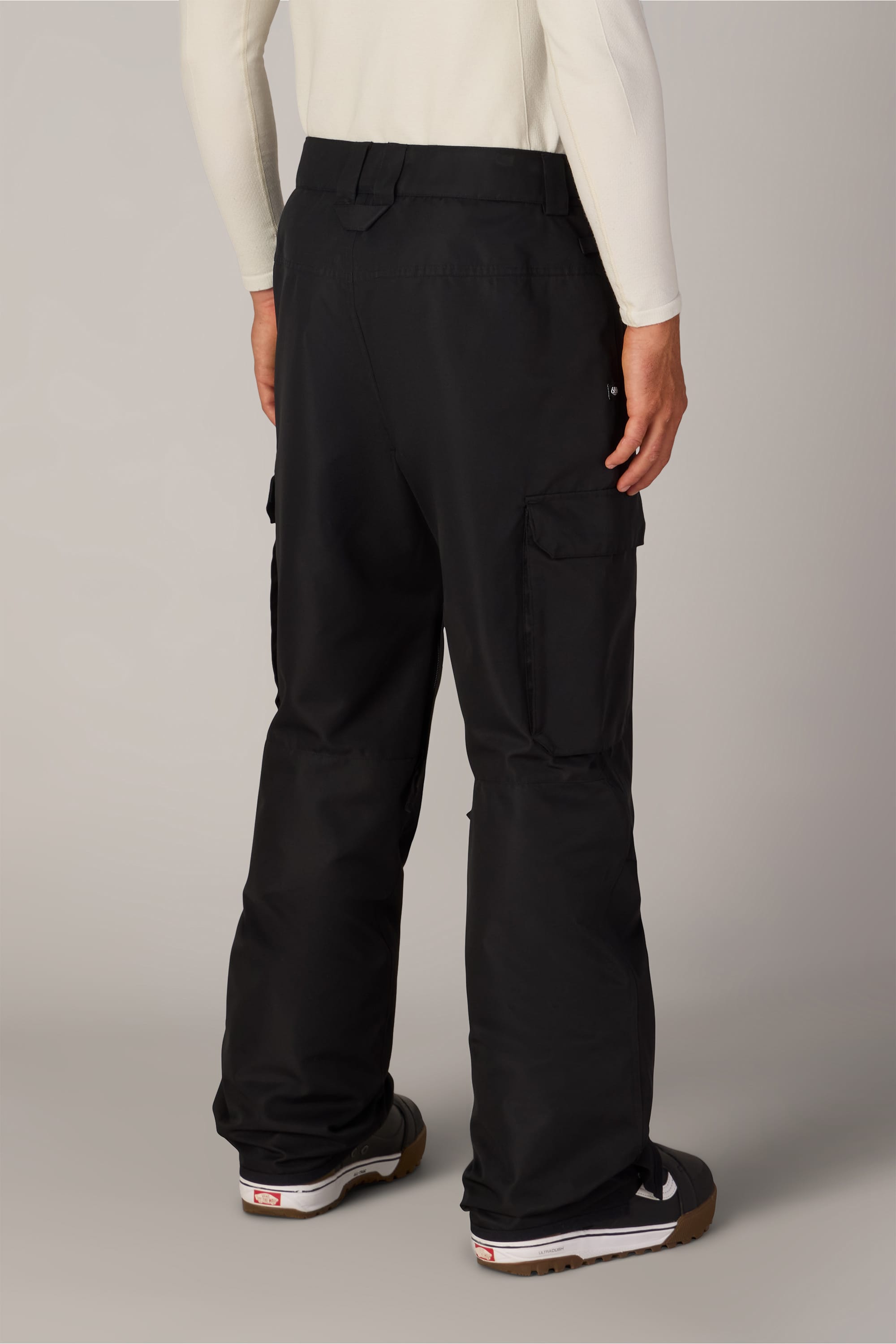 A person wearing the 686 Men’s 686 Nightline Cargo Pant in black and a white long-sleeve shirt, viewed from the back. The pants by 686 feature side pockets and are crafted from waterproof woven fabric, combining practicality with style. Black sneakers with white soles complete the outfit, set against a plain, neutral background.