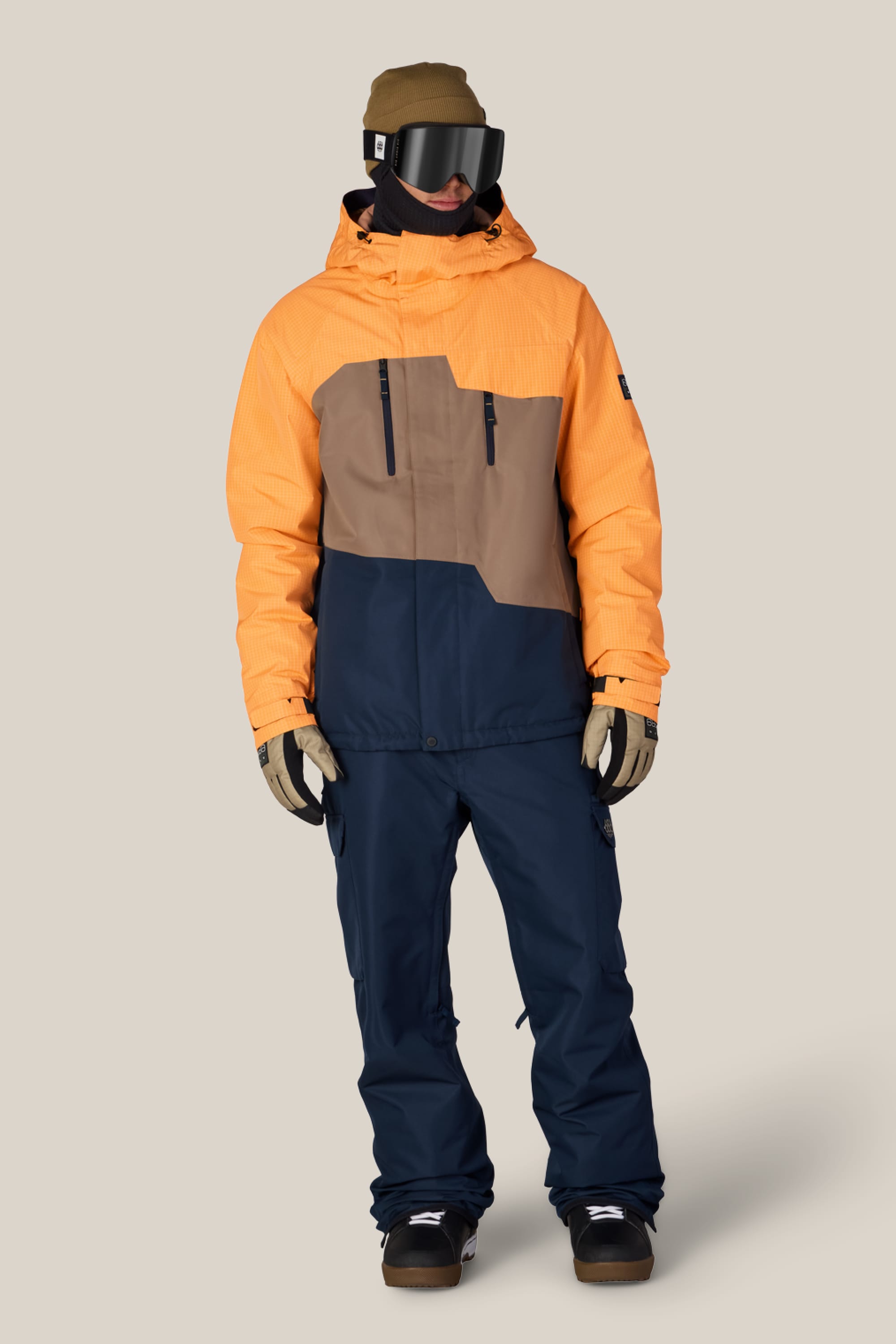 Person wearing a color-blocked orange, tan, and navy blue ski jacket paired with 686 Men’s Nightline Cargo Pant in navy polyester waterproof fabric by 686, along with tan gloves, a brown hat, black goggles, and a black face covering, standing against a plain beige background.