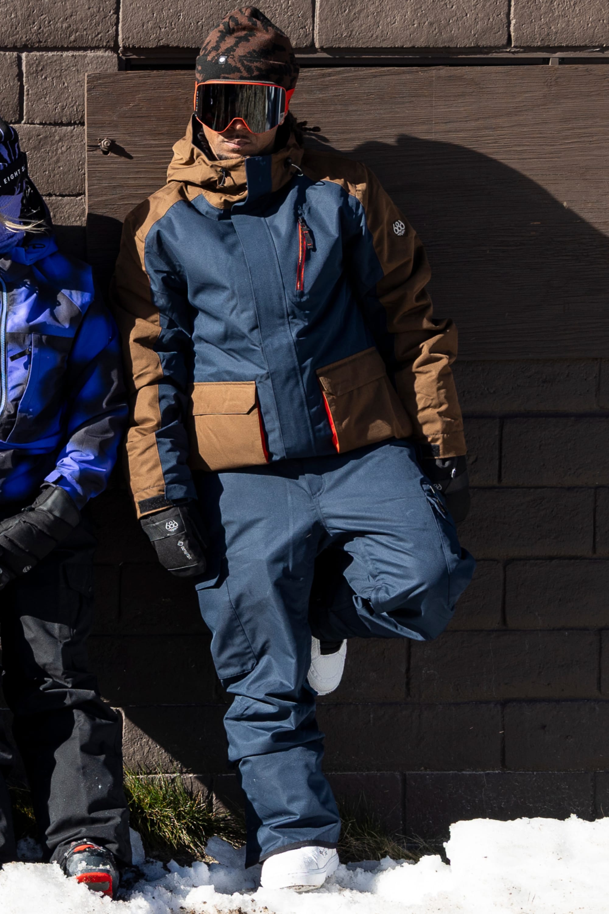 A person in a blue and brown snowsuit with white boots, black gloves, and large reflective ski goggles stands against a brick wall with one knee bent in a snowy area. The look is completed with the 686 Men’s Nightline Cargo Pant by 686, designed for winter performance. Another individual wearing blue winter gear is partly visible.
