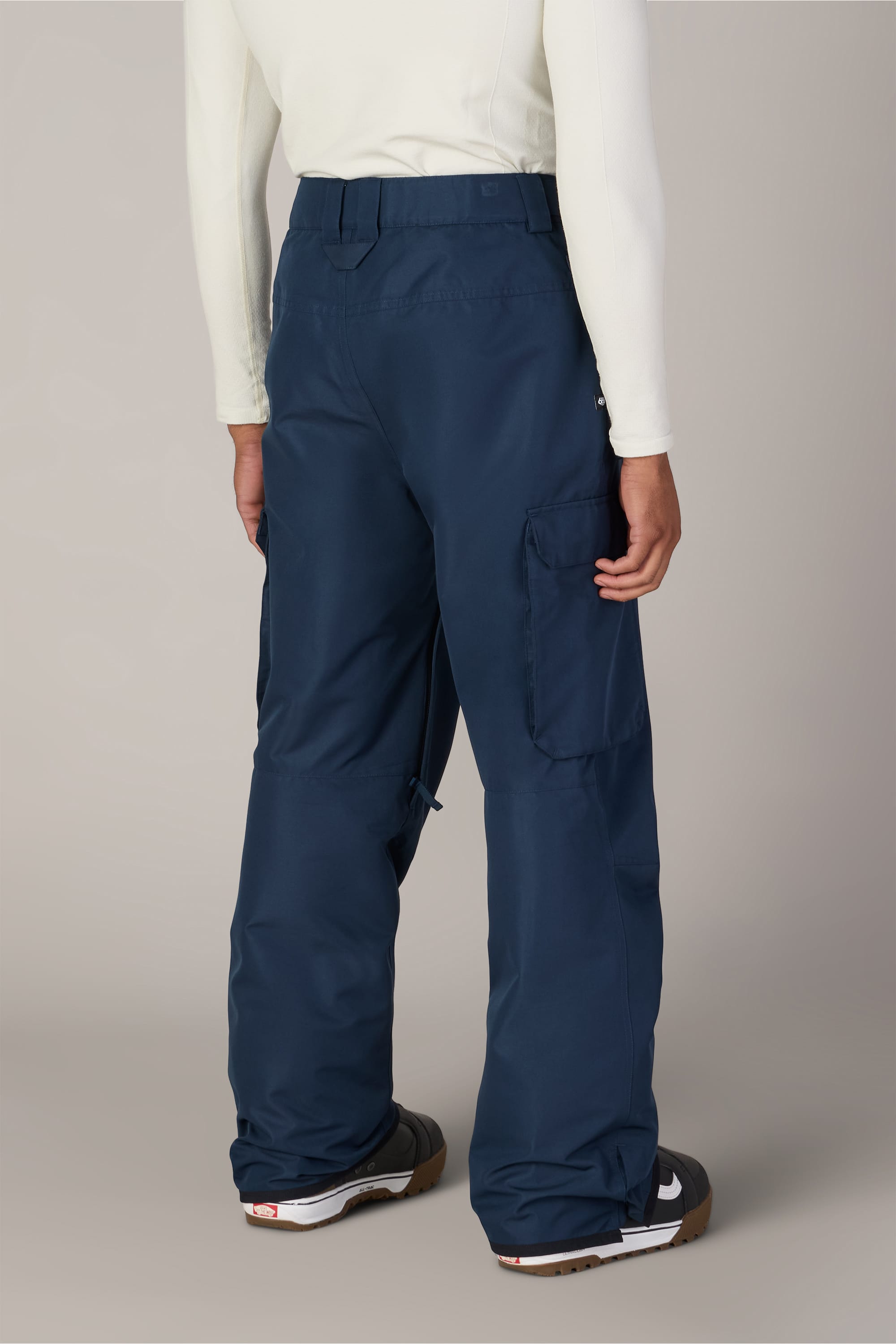 A person wearing a white long-sleeve shirt, black shoes, and 686 Men’s 686 Nightline Cargo Pant in blue by 686, shown from the back, standing against a plain light background.