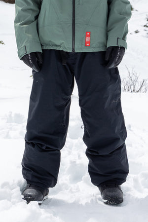 A person standing in the snow is wearing a green jacket, black 686 Dojo® GORE-TEX® Shell Pants, black gloves, and black boots. The snow is white and the background features more snowy ground dotted with some vegetation.