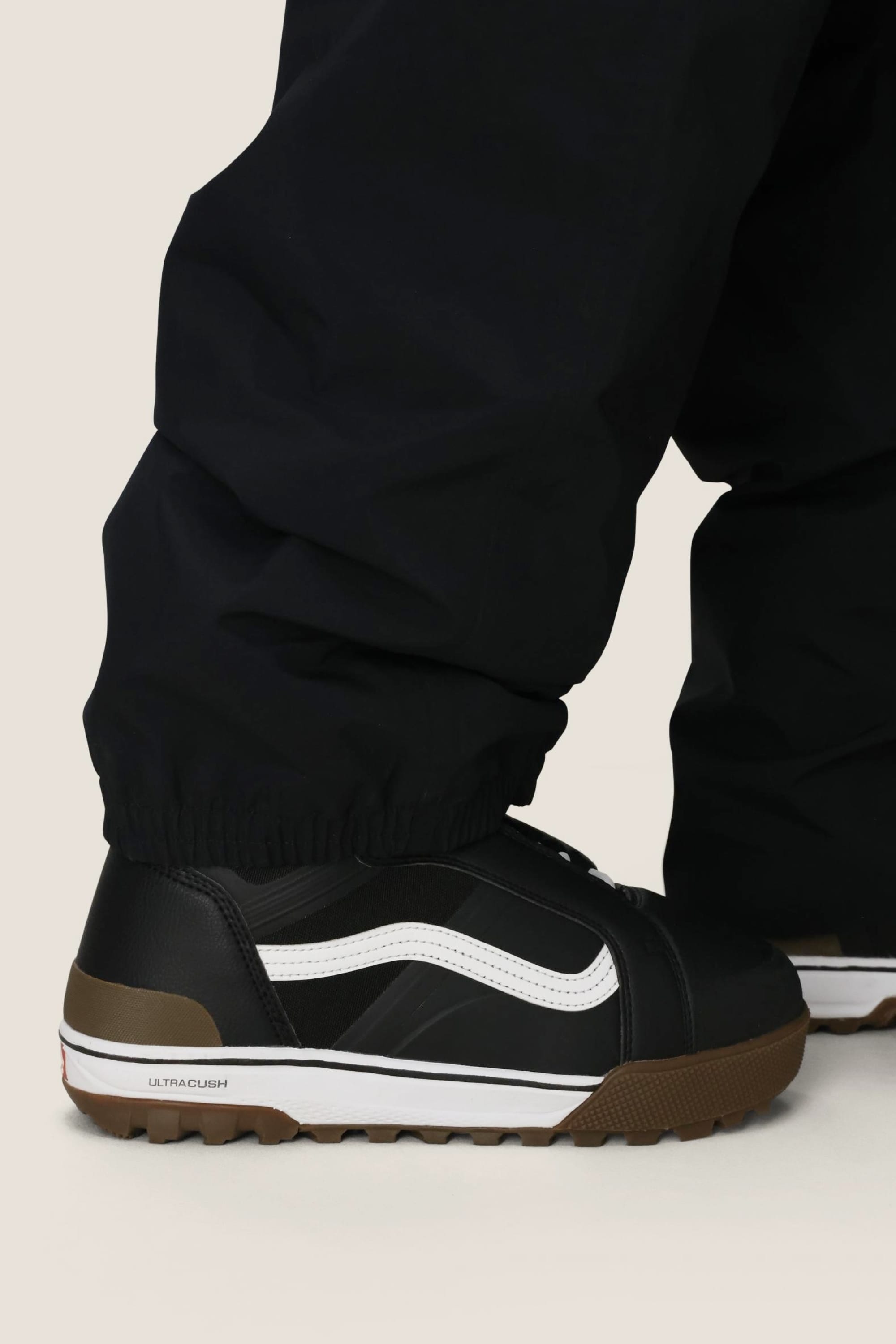A person wearing the 686 Men’s 686 Dojo® GORE-TEX® Shell Pant by 686 and black Vans sneakers with white stripes and brown soles stands on a light background.