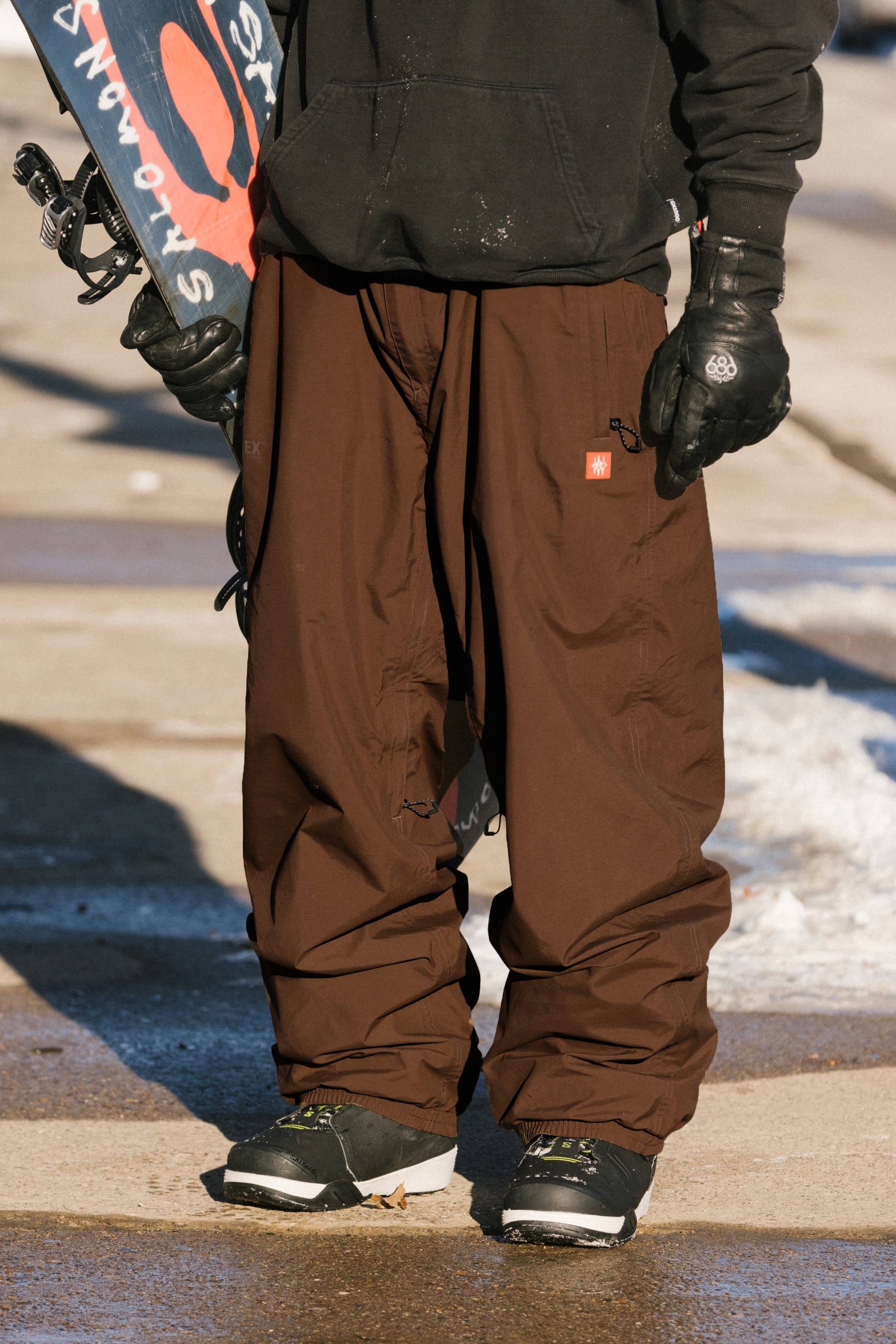 A person wearing the 686 Men’s Dojo® GORE-TEX® Shell Pant by 686, along with a black hoodie, gloves, and sneakers, stands on a sidewalk holding a snowboard. Some snow is visible on the ground.
