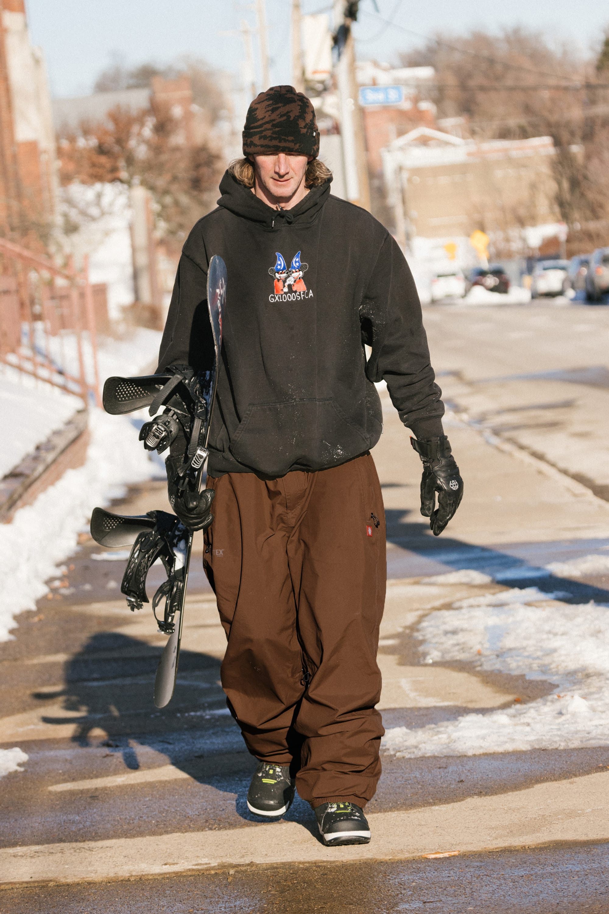 A person wearing the 686 Men’s 686 Dojo® GORE-TEX® Shell Pant, a black hoodie, and a camo hat walks down a snowy street holding a snowboard in one hand.