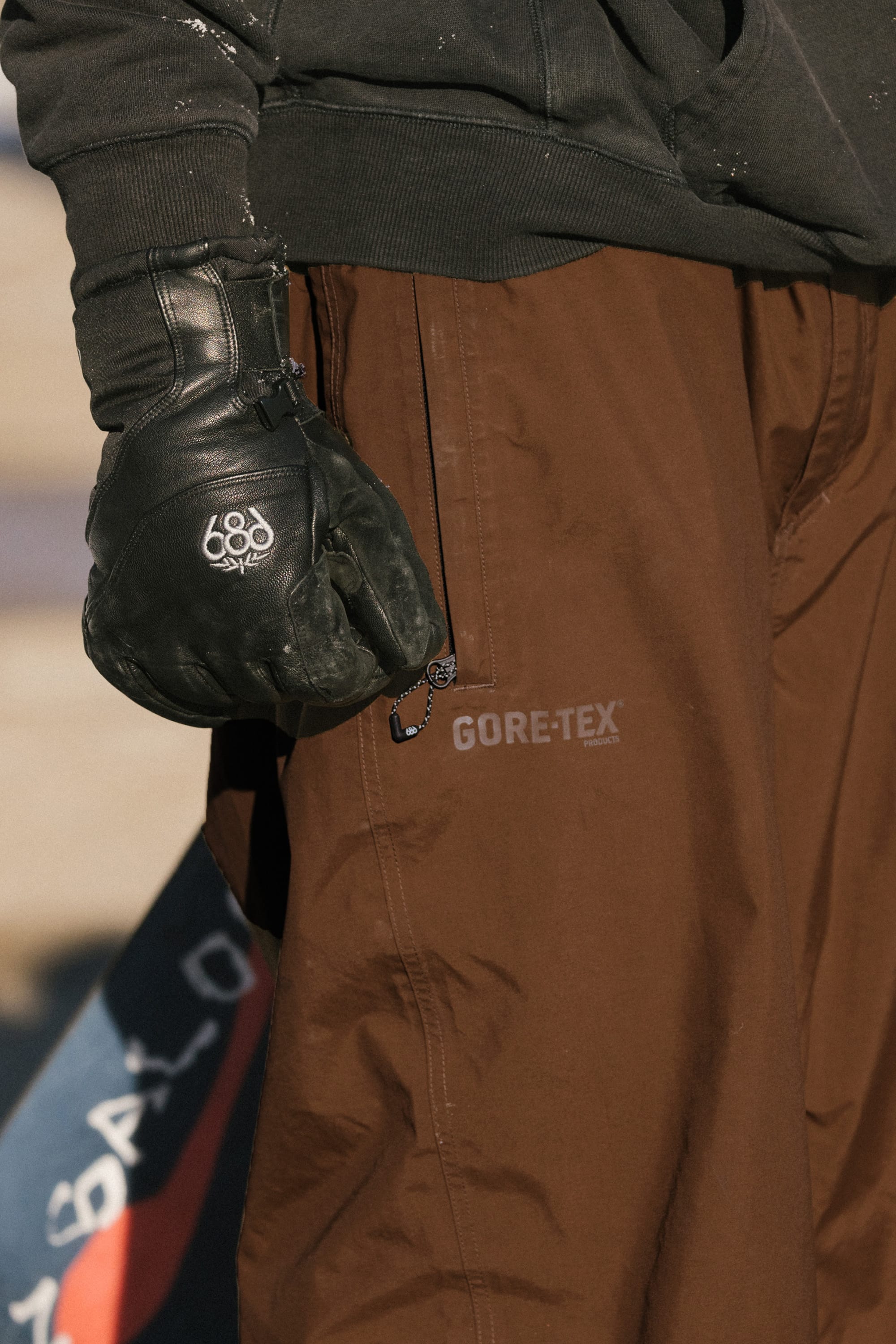 A person wearing black gloves and the 686 Men’s 686 Dojo® GORE-TEX® Shell Pant stands outdoors, holding a dark object. Only their glove, pants, and part of a dark jacket are visible.