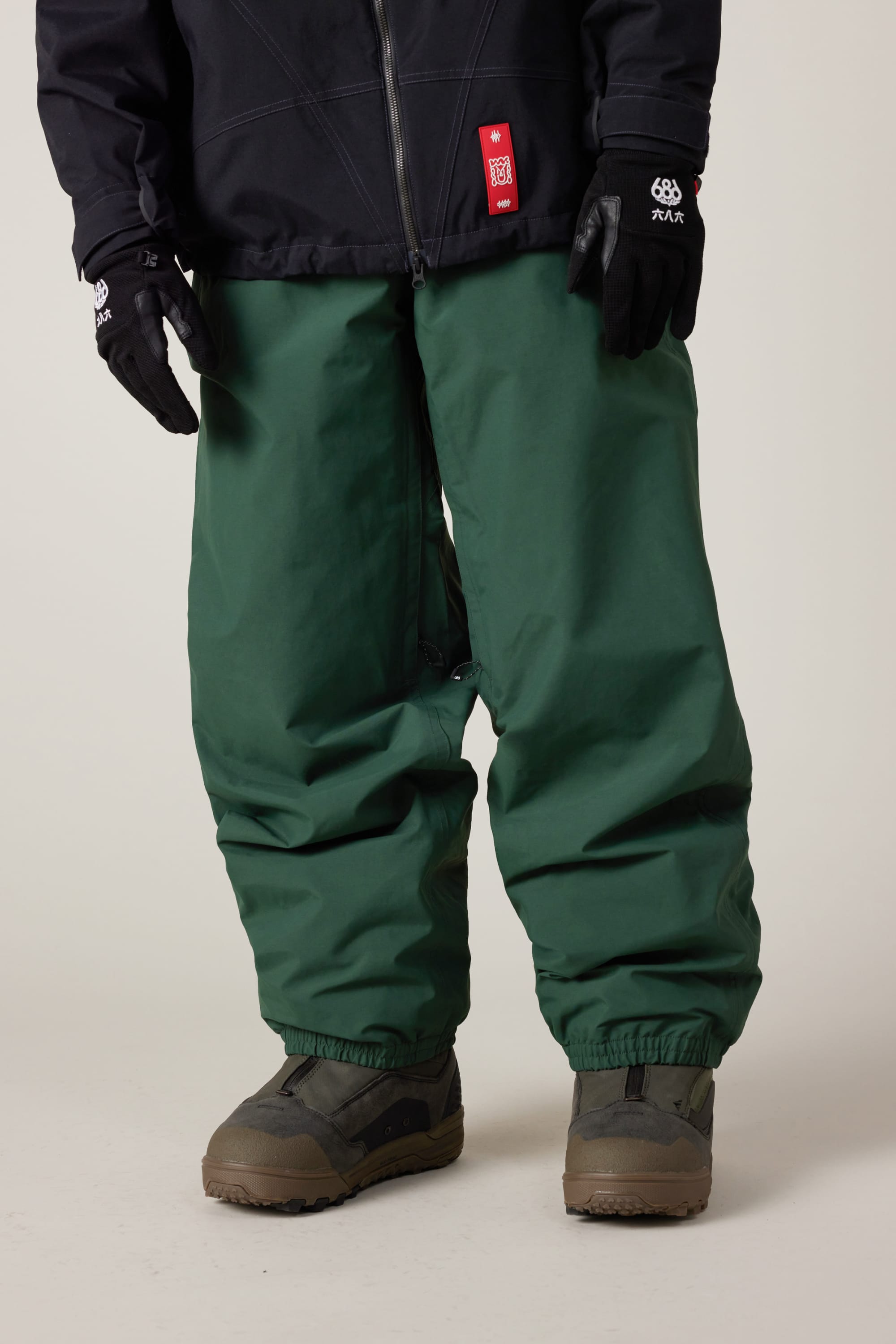 A person wearing 686 Men’s 686 Dojo® GORE-TEX® Shell Pant by 686 in green, a black jacket with a red tag, black gloves, and brown outdoor boots stands against a plain background.