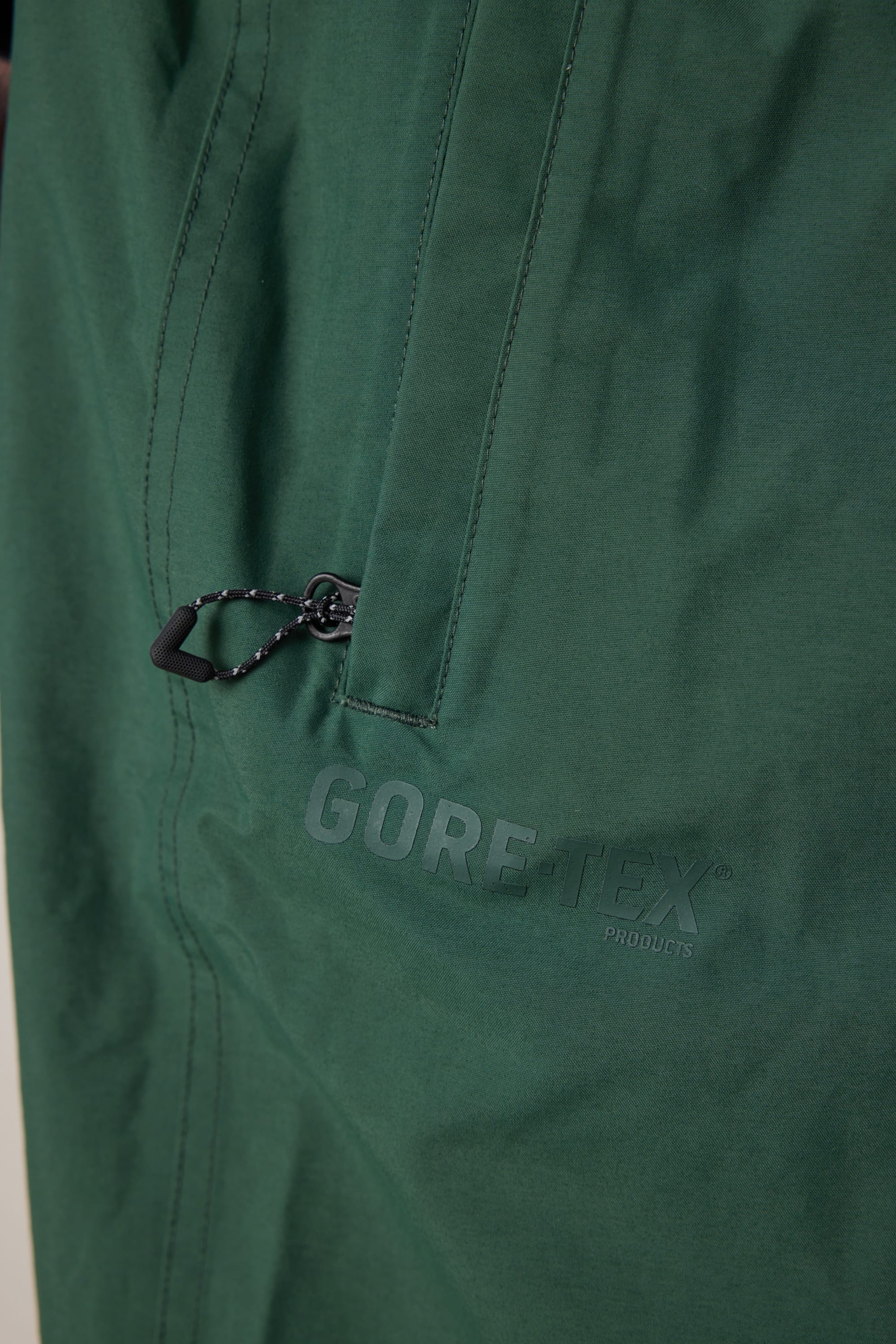 Close-up of the green fabric on the 686 Men’s 686 Dojo® GORE-TEX® Shell Pant, featuring a zippered pocket with a black pull cord and the GORE-TEX PRODUCTS logo printed on it—details that emphasize the technical, waterproof design from 686.