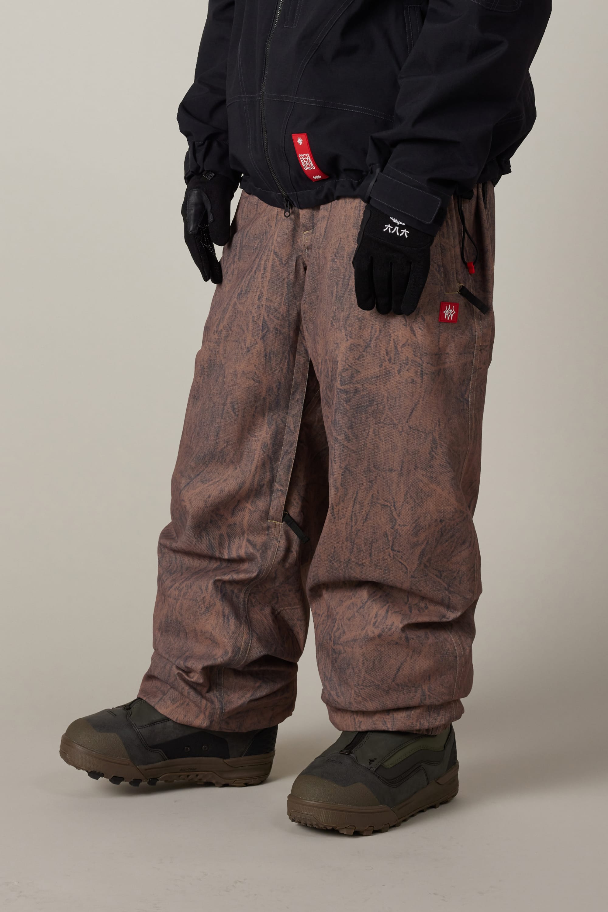 Person wearing baggy brown 686 Men’s 686 Dojo® Shell Pant, black gloves, black jacket, and olive green winter boots, standing against a plain background.
