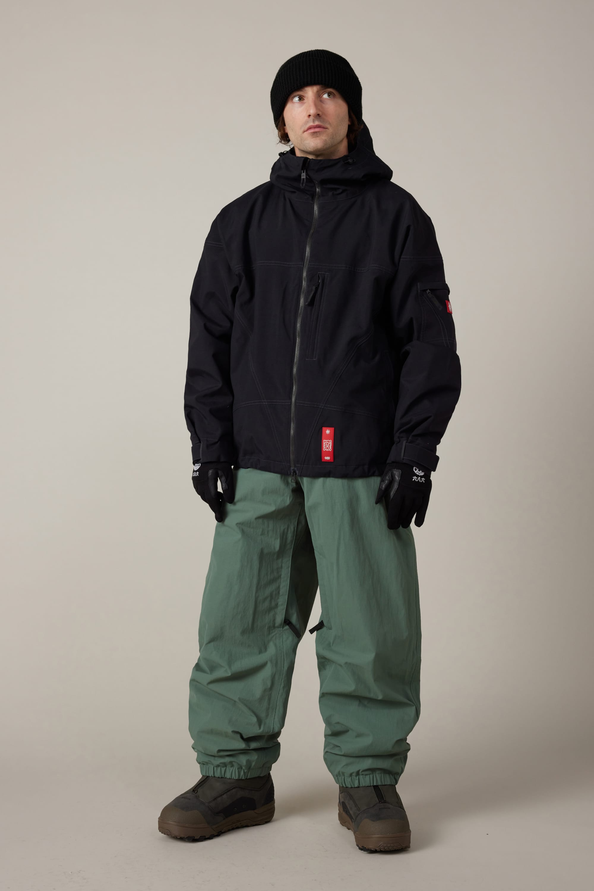 A person wearing a black beanie, black zip-up jacket, 686 Men’s 686 Dojo® Shell Pant by 686 in green, black gloves, and brown boots stands facing forward against a plain light background.