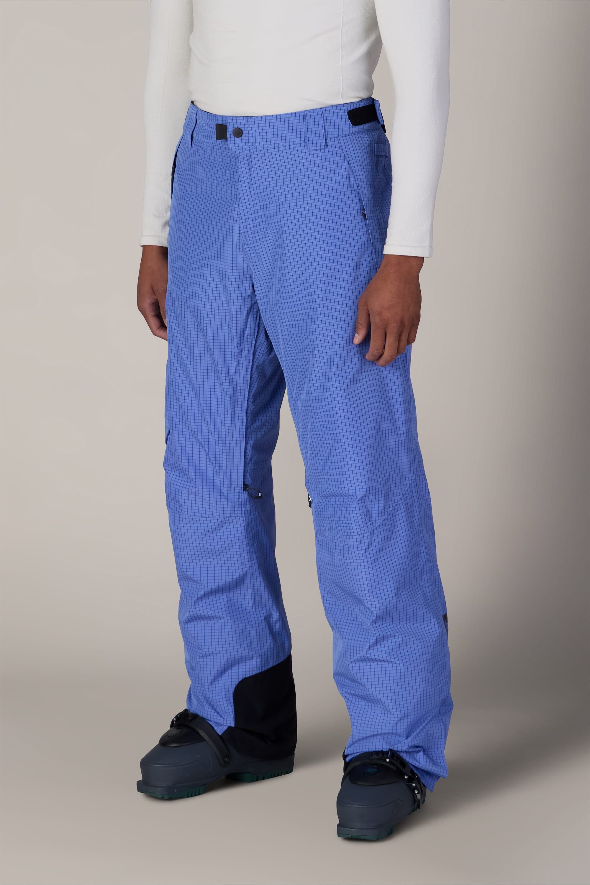 A person wearing the 686 Men’s Hydra™ Thermagraph® Pant in a light blue checkered pattern, paired with a white long-sleeve shirt and black snow boots, stands against a neutral background.