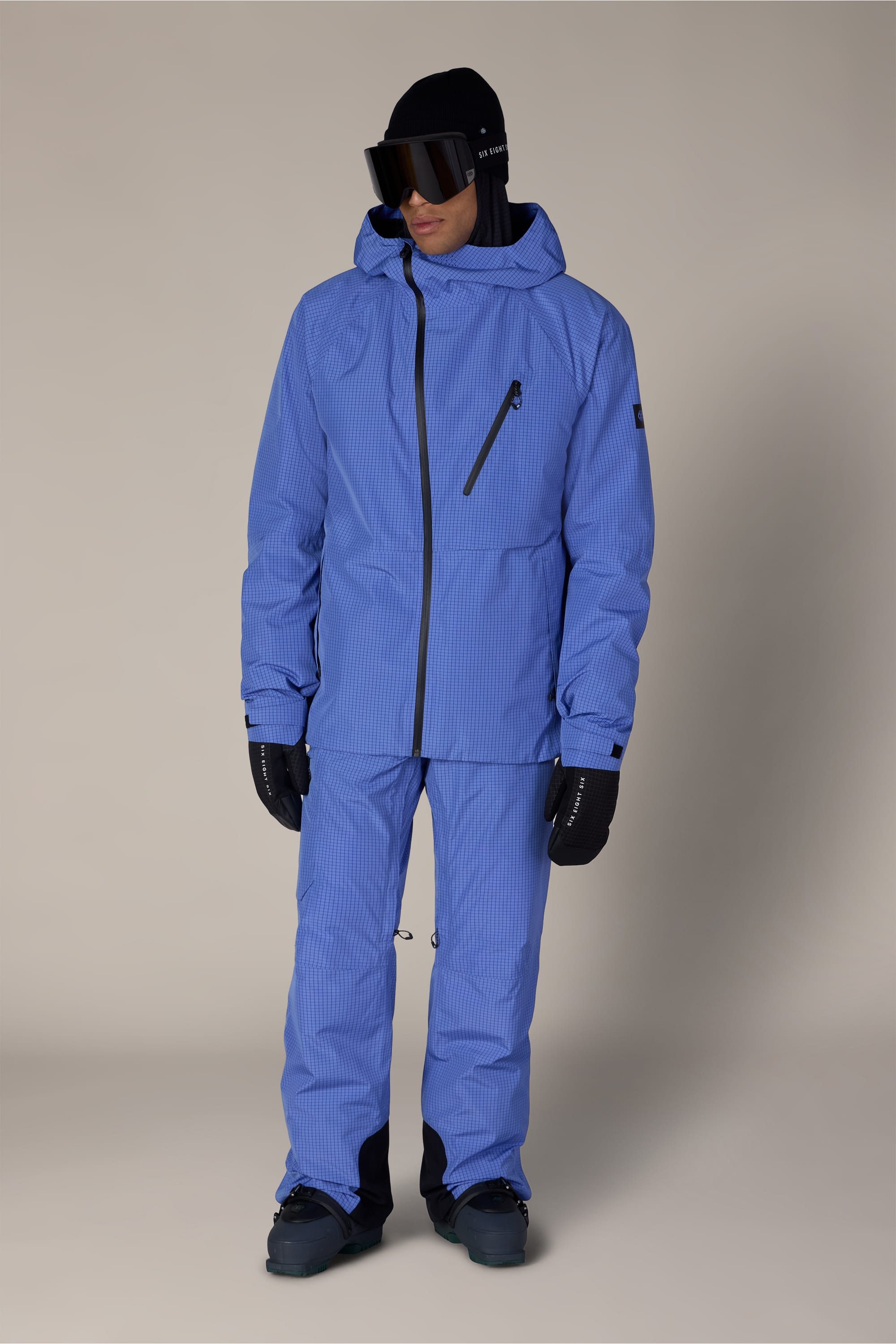 A person wearing a matching light blue ski jacket and 686 Men’s Hydra™ Thermagraph® Pant by 686, along with black gloves, black boots, and dark ski goggles, stands against a plain beige background.