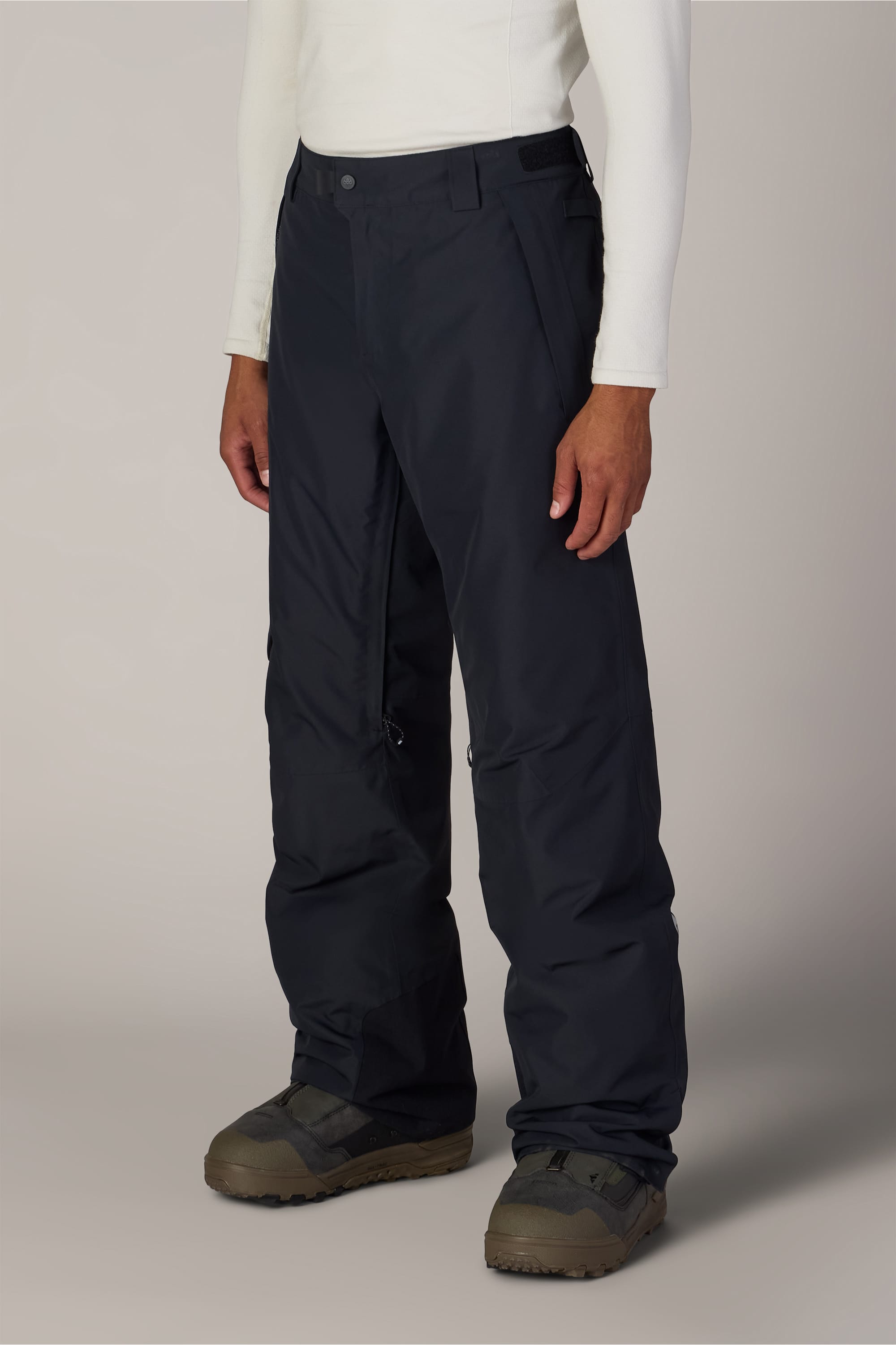 A person is shown from the waist down wearing black 686 Men’s Hydra™ Thermagraph® Pants, white long-sleeve shirt, and gray winter boots against a plain light background.