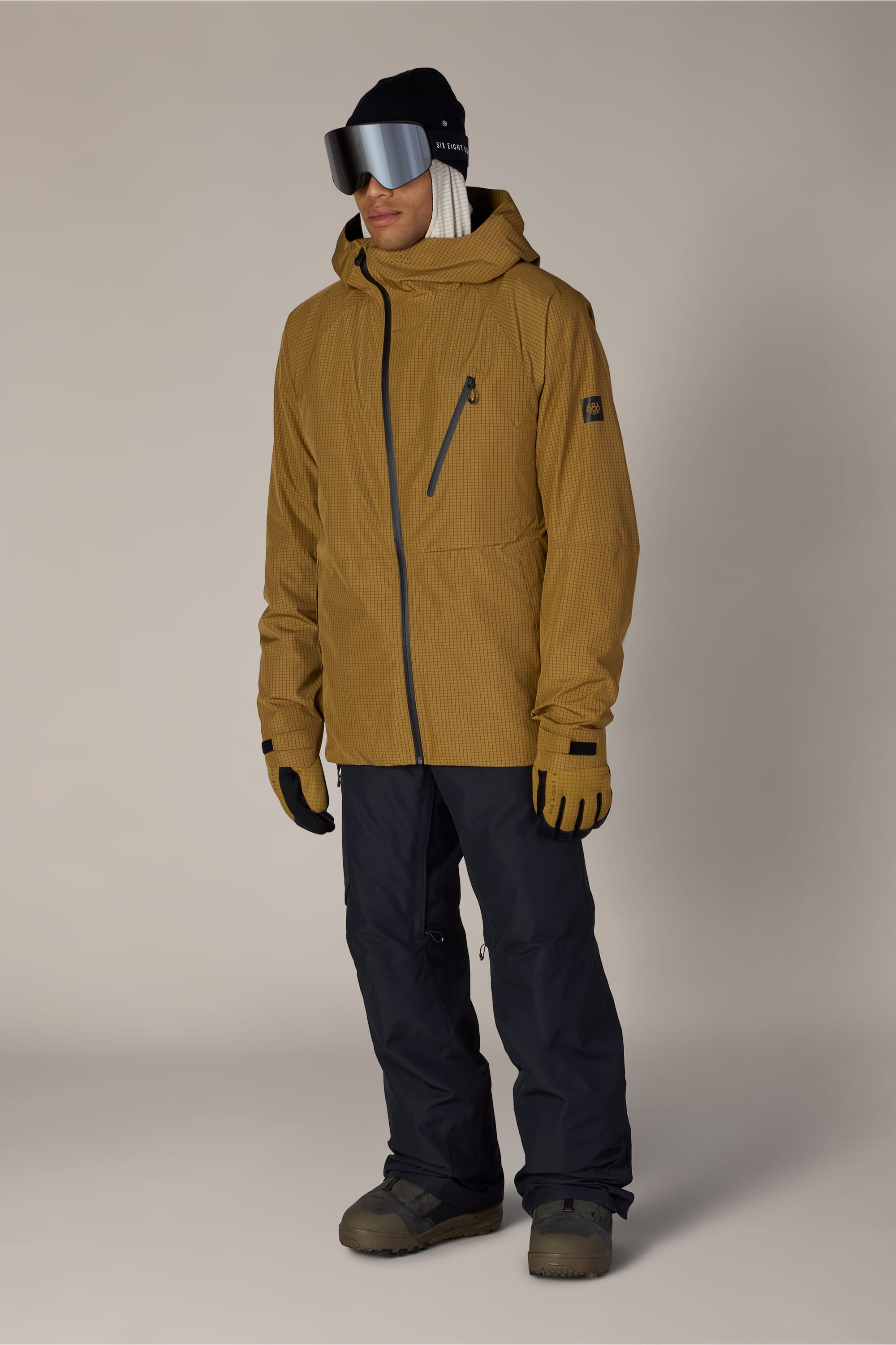 A person wearing black 686 Men’s Hydra™ Thermagraph® Pants, a mustard yellow ski jacket, gloves, black ski goggles, a white beanie, and snow boots stands against a plain light background.