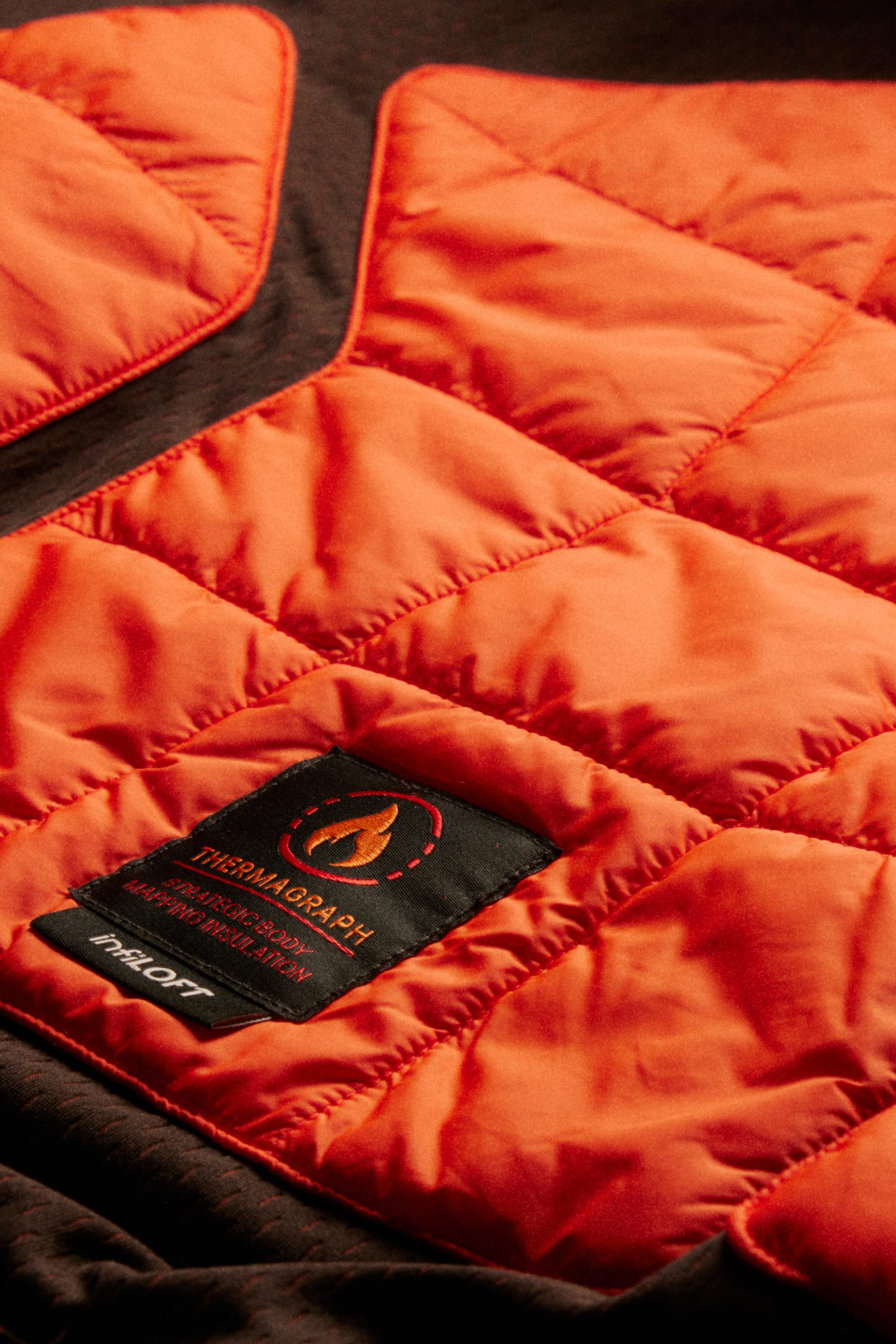 Close-up of an orange, quilted jacket section features a black label reading “THERMAGRAPH” with a flame icon, showcasing the insulation and design details—ideal for pairing with the 686 Men’s Hydra™ Thermagraph® Pant from 686 for dependable warmth and protection in cold, wet conditions.