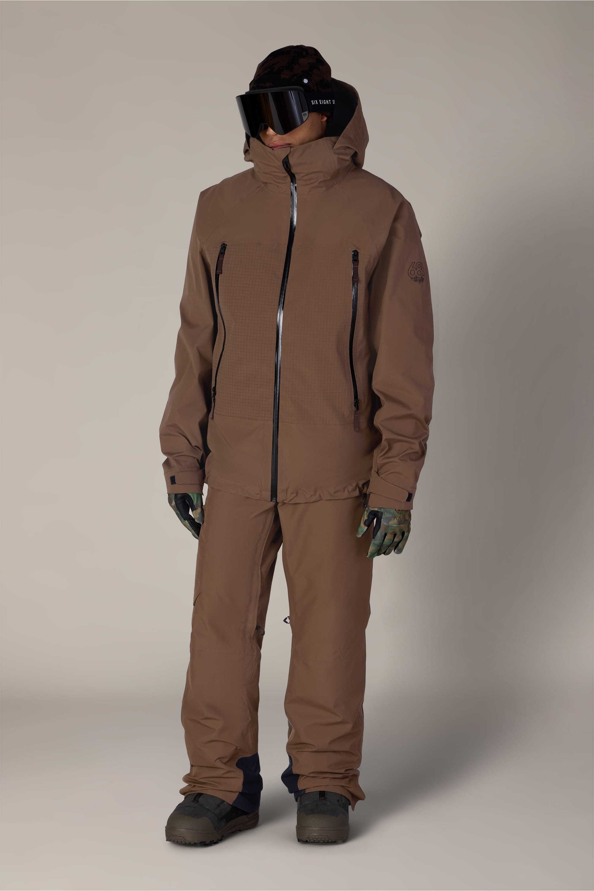 A person dressed in the 686 Men’s Hydra™ Thermagraph® Pant by 686, paired with a brown hooded jacket, dark gloves, black ski goggles, and boots stands against a plain light background. Their face is fully covered, and the outfit is suitable for outdoor or winter sports activities.