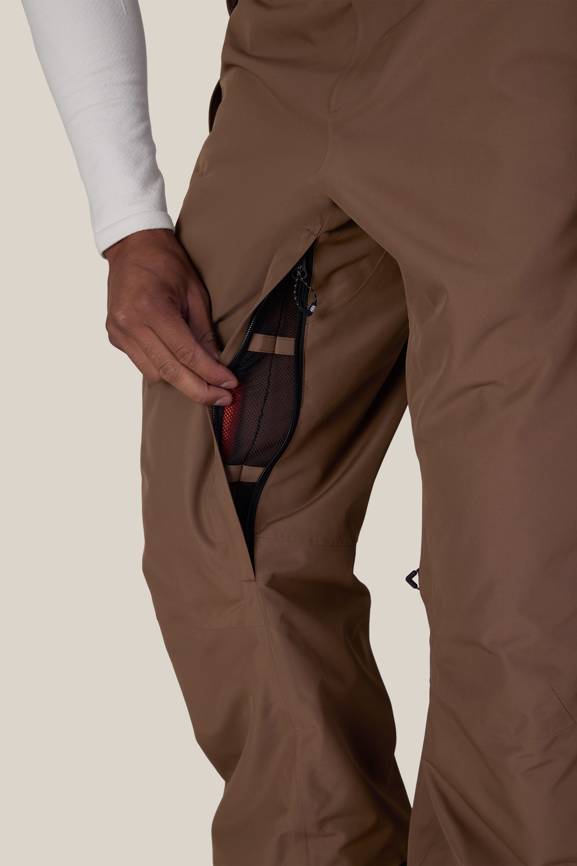 A person opens a mesh-lined zipper vent on the thigh of the brown 686 Men’s Hydra™ Thermagraph® Pant, revealing a red inner pocket.