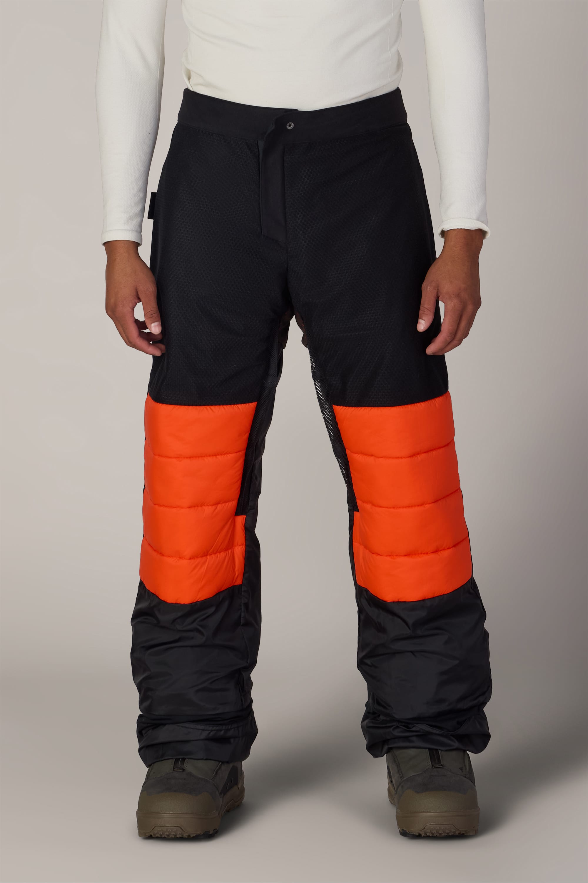 A person wearing 686 Men’s Hydra™ Thermagraph® Pant by 686, featuring black polyester fabric with bright orange padded panels on the thighs, paired with a white long-sleeve shirt and brown shoes, standing against a plain background.