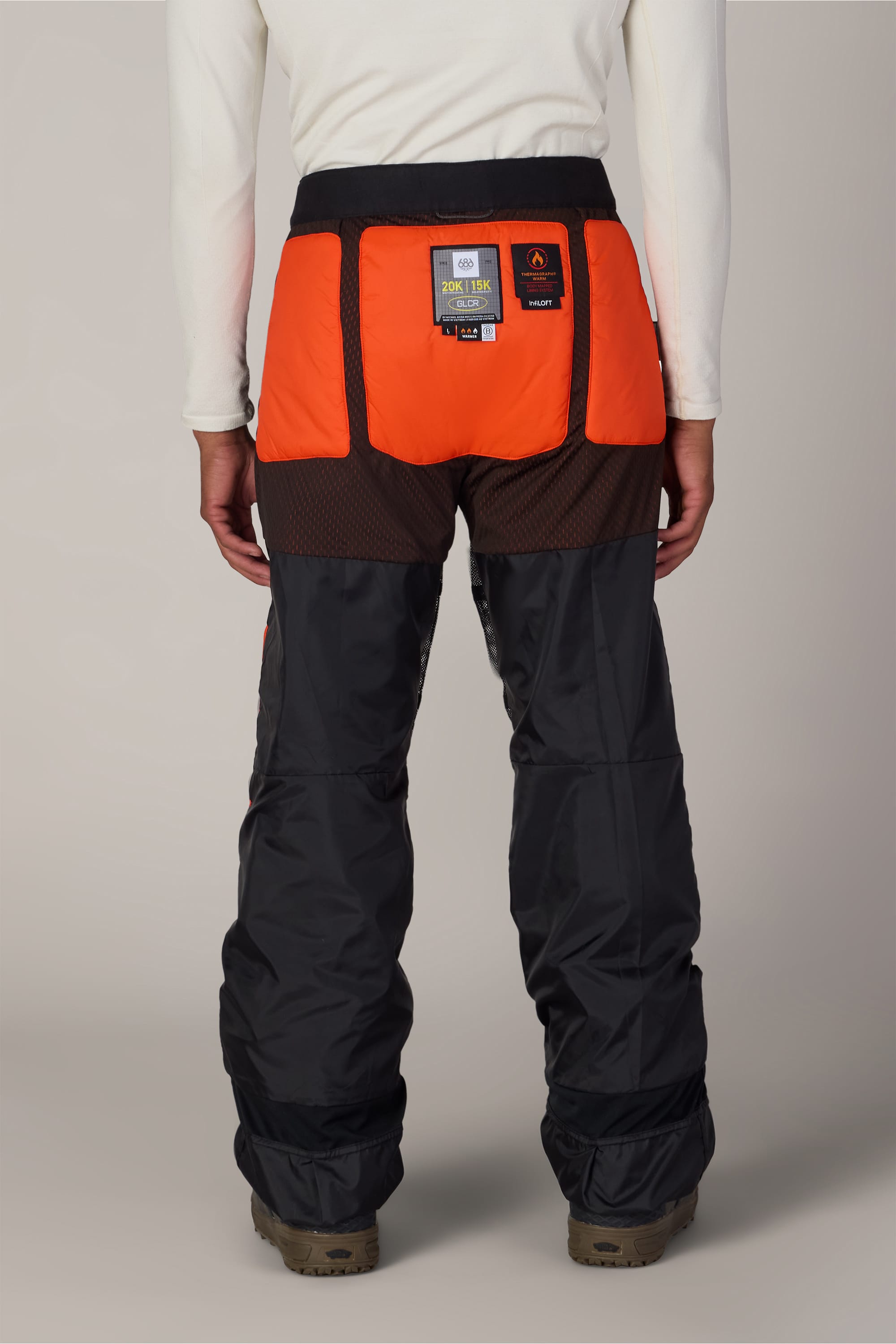 A person stands facing away, wearing 686 Men’s Hydra™ Thermagraph® Pant by 686—black and orange insulated nylon pants with mesh panels, featuring a visible patch and tag on the back waistband. They are also wearing brown boots, set against a plain light-colored background.