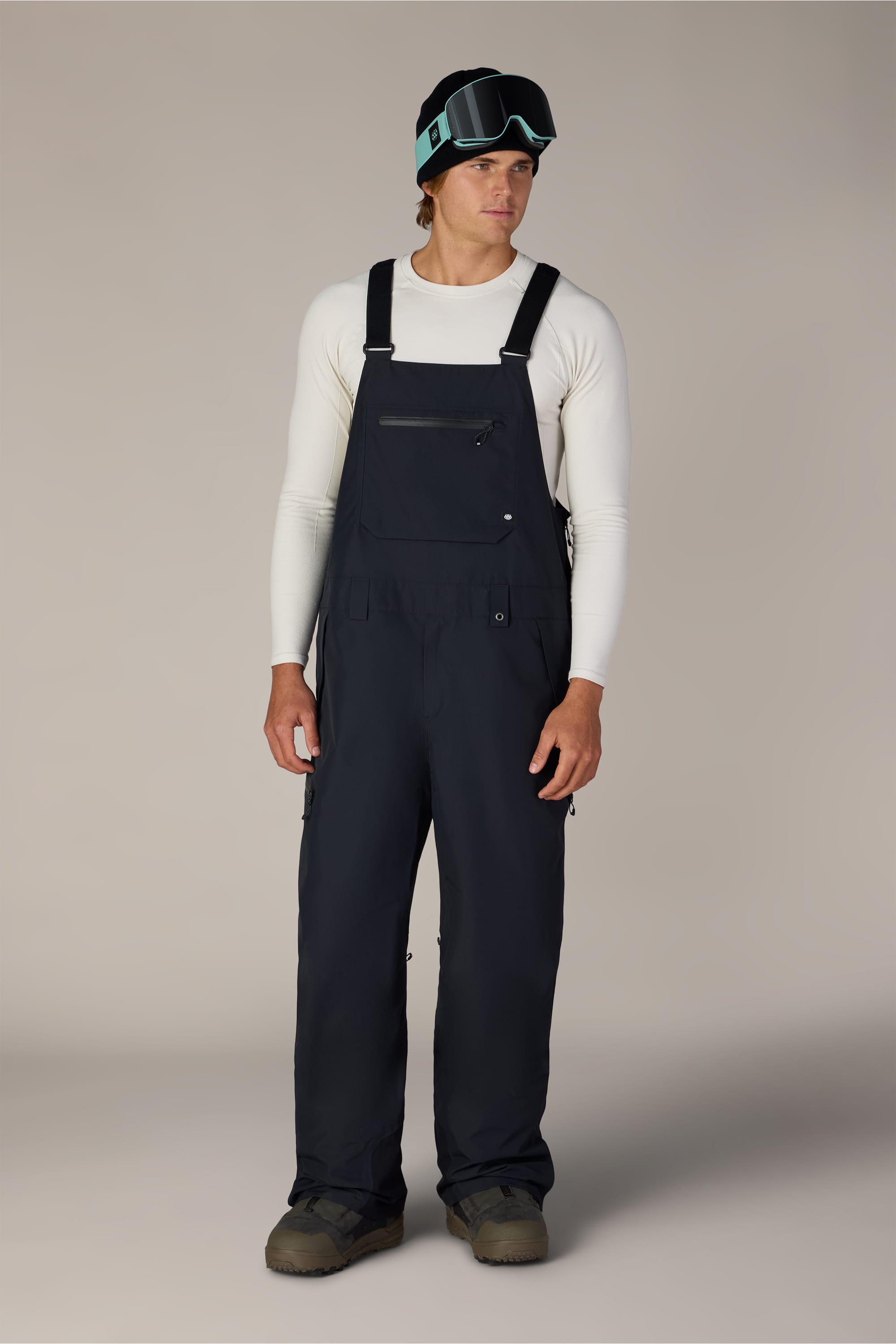 A person wearing the 686 Men’s Lockup™ Shell Bib by 686, along with a white long-sleeve shirt, dark boots, and a black helmet with ski goggles, stands facing forward against a plain background.