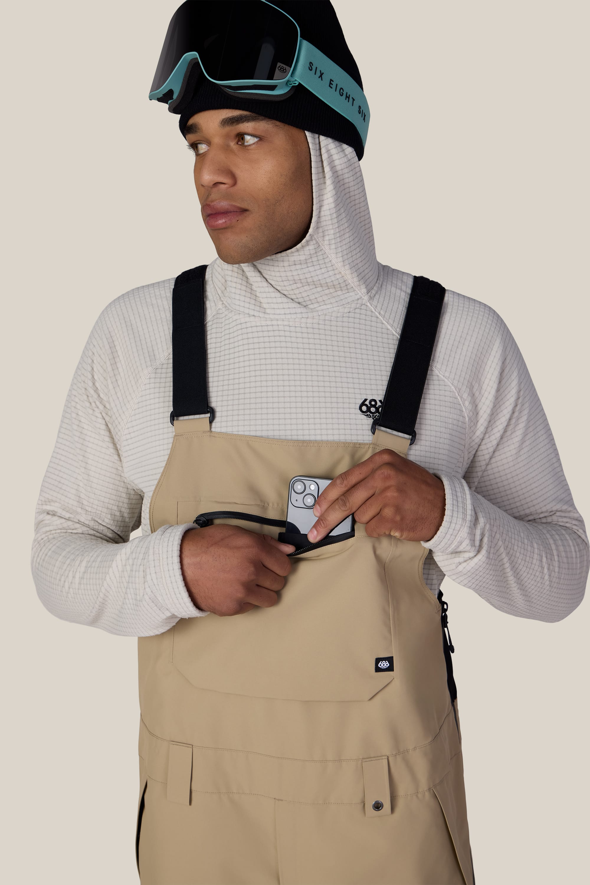 A man wearing the 686 Men’s Lockup™ Shell Bib in beige and a white hoodie, with ski goggles resting on his head, places a smartphone into the bib’s chest pocket while looking to the side against a plain background.