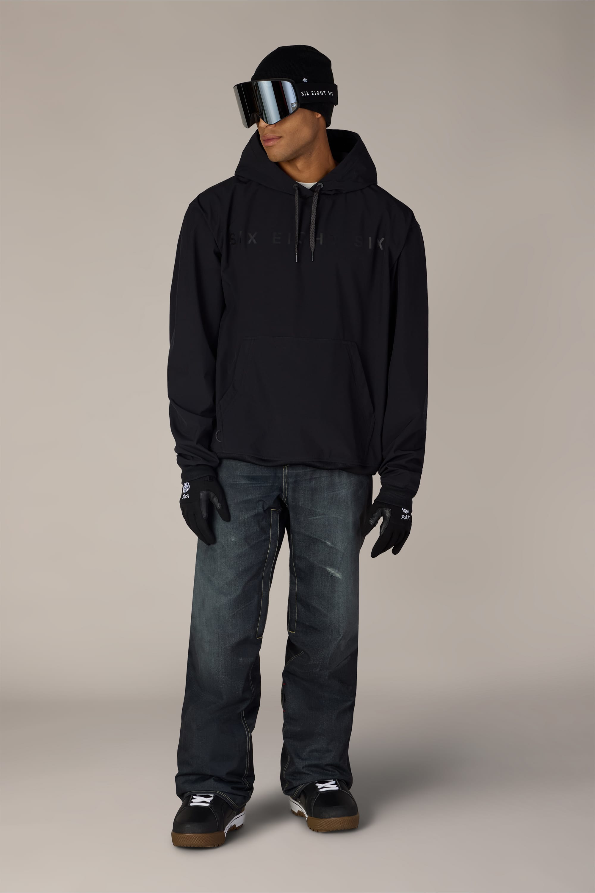 A person wearing a black hoodie, black gloves, the 686 Men’s Deconstructed™ Denim Insulated Pant by 686, black sneakers, a black beanie, and large reflective ski goggles stands against a plain background, looking to the side.