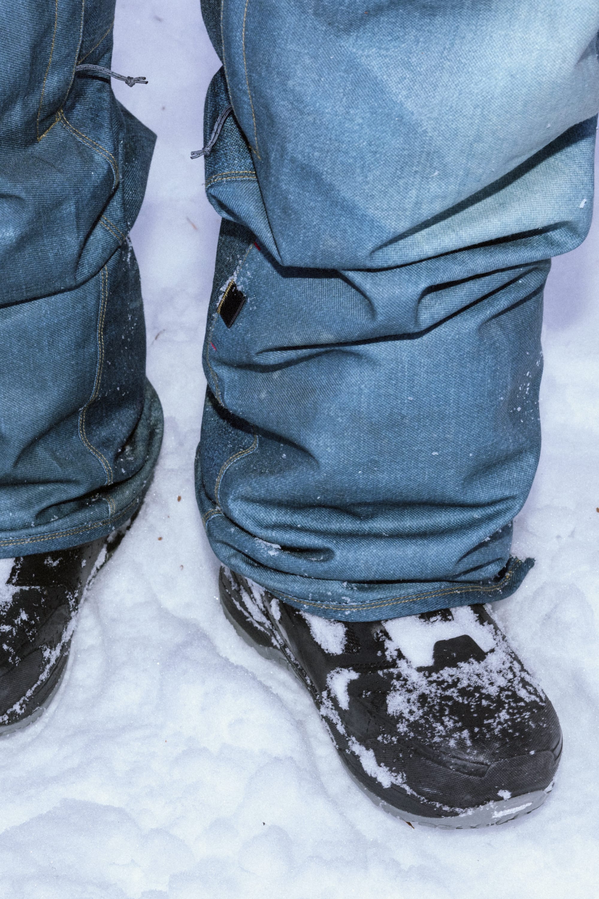 A close-up of someone wearing the 686 Men’s Deconstructed™ Denim Insulated Pant and black snow boots standing on snow, with bits of snow on the waterproof pants and boots.