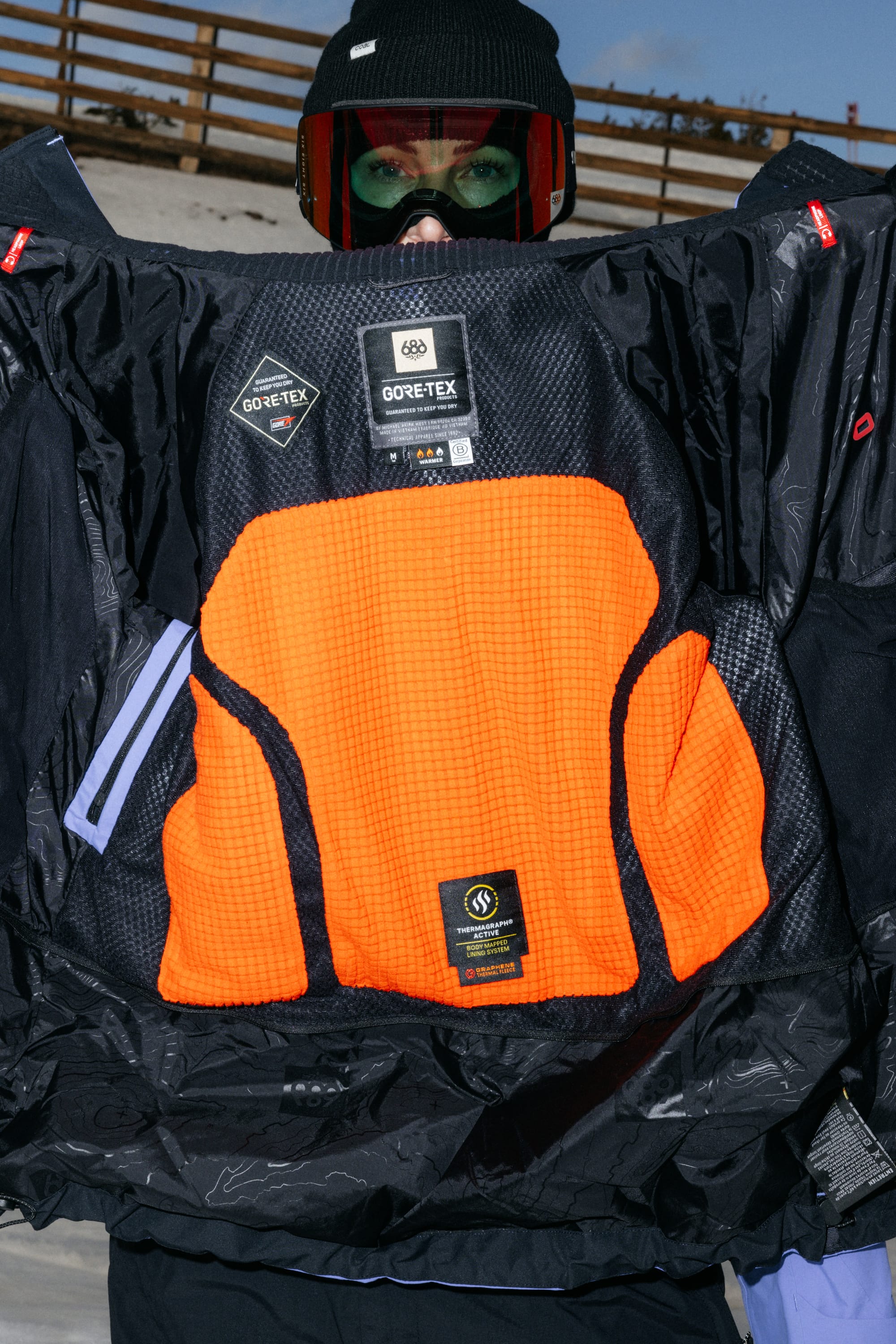 A person in winter gear holds open a 686 Women’s GORE-TEX® Scheme Thermagraph® Jacket, revealing its bright orange and black GORE-TEX interior lining along with several visible brand and care labels.