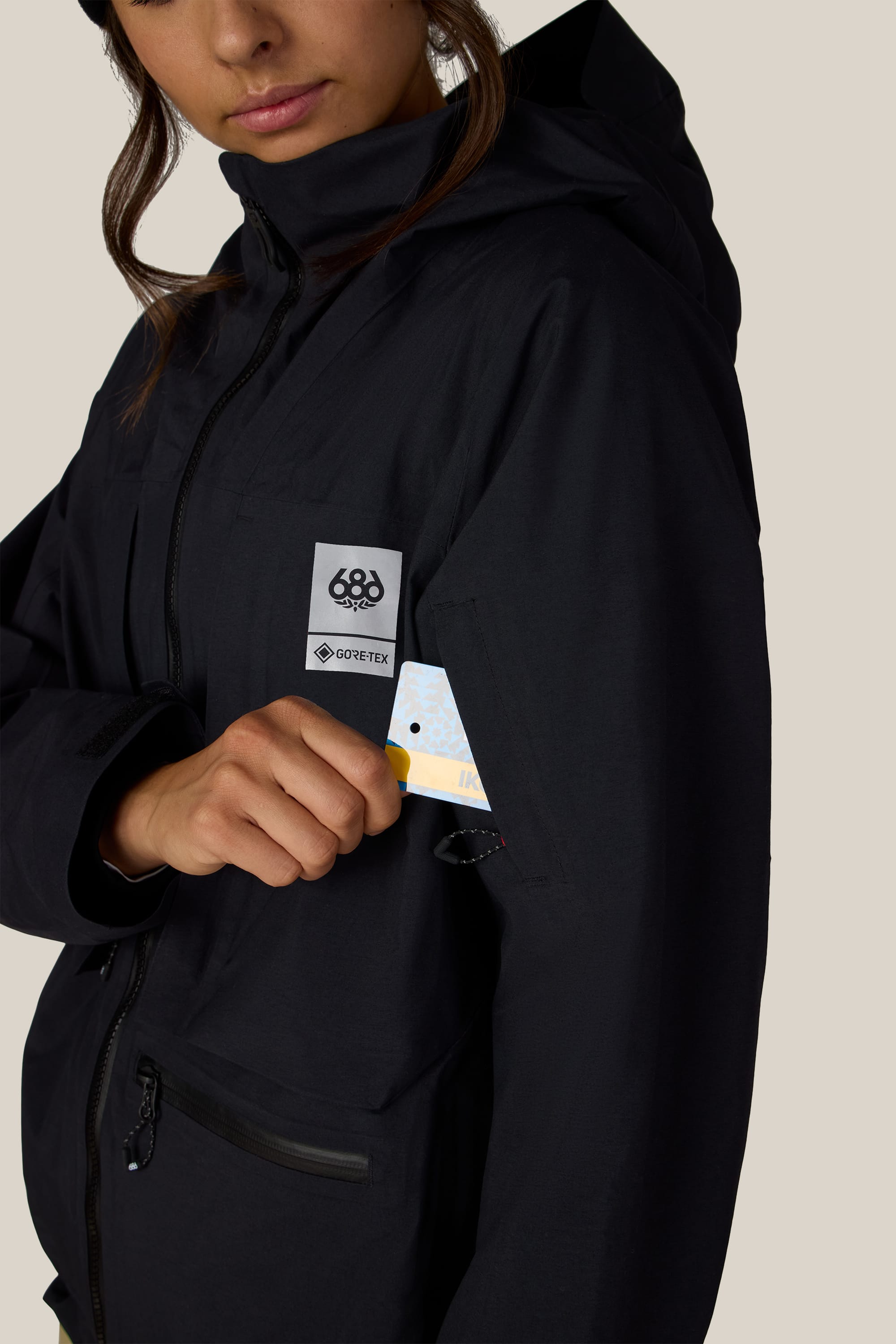 A person wearing the 686 Women’s GORE-TEX® Scheme Thermagraph® Jacket in black pulls a colorful card from a zippered arm pocket. The jacket features a Gore-Tex patch and the 686 logo on the chest.
