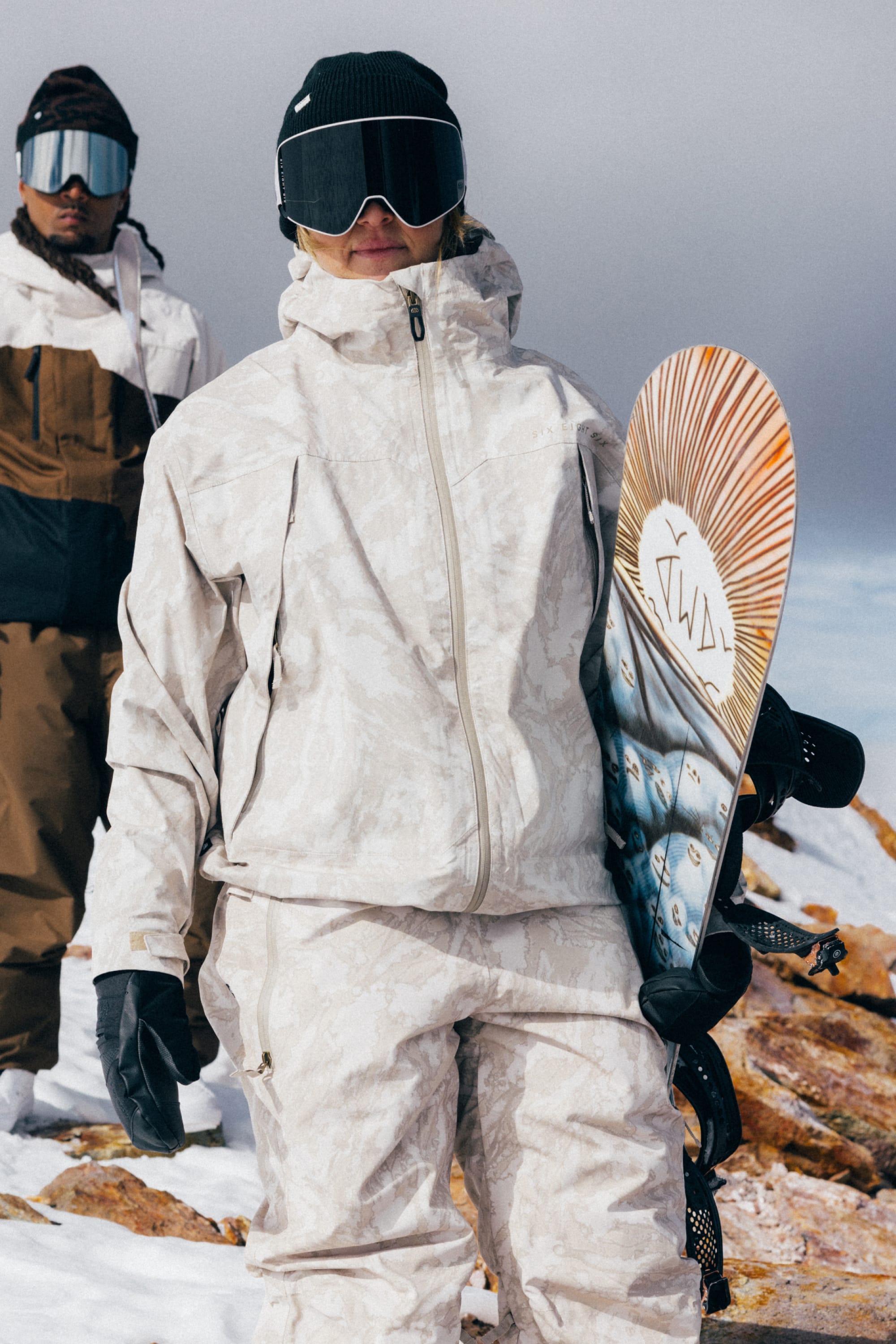Two people in winter gear stand on a snowy mountain. One holds a snowboard with a colorful design. Both wear ski goggles, gloves, and jackets—one showcasing the 686 Women’s SMARTY® 3-in-1 Framework™ 3L Jacket from 686 with a sleek finish—while rocky terrain and a cloudy sky form the background.