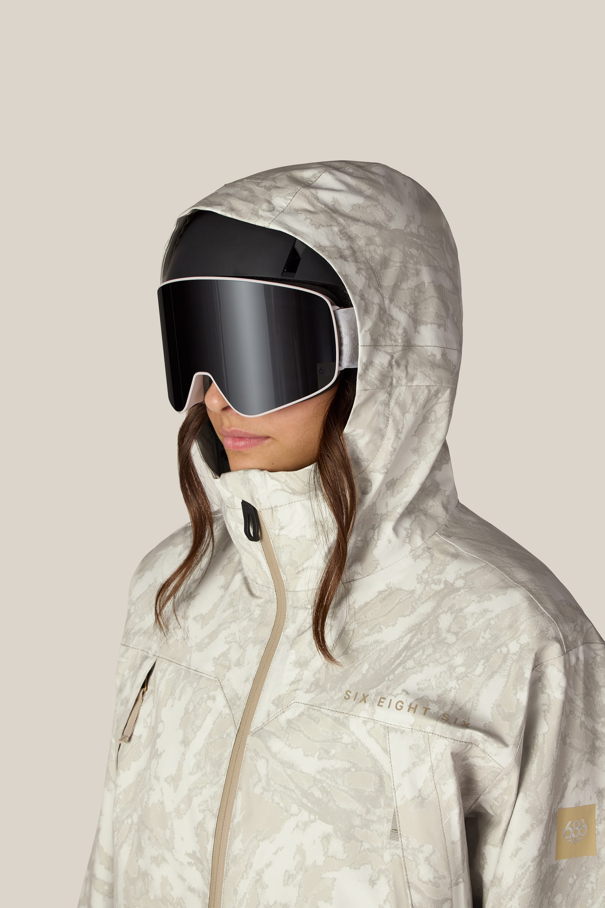 A person is wearing the 686 Women’s SMARTY® 3-in-1 Framework™ 3L Jacket by 686—a white and gray patterned women’s woven jacket with a hood—along with large black ski goggles, facing slightly to the left against a plain beige background.