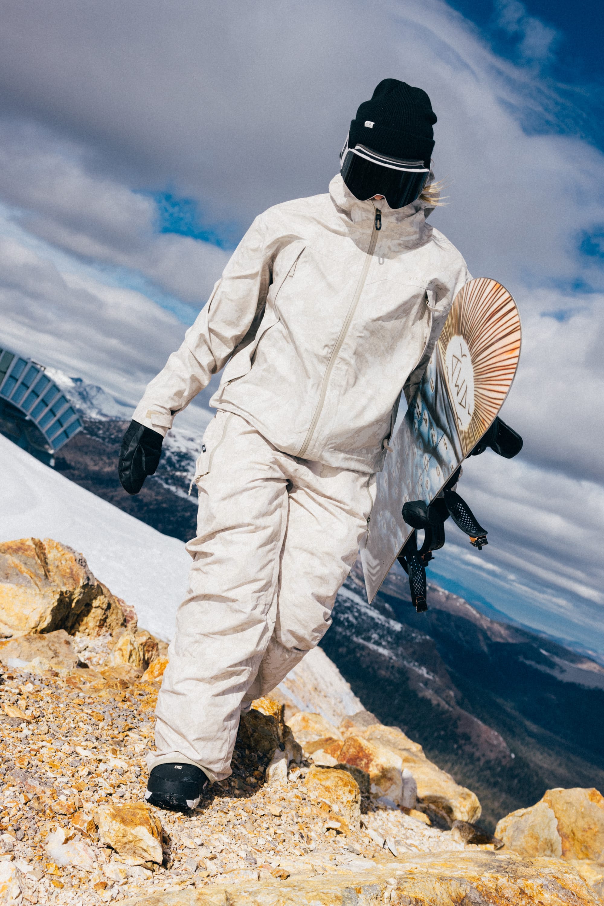 Wearing the 686 Women’s SMARTY® 3-in-1 Framework™ 3L Jacket, a snowboarder in black gloves and a black beanie walks across rocky terrain with a snowboard under her arm. Behind her, snowy mountains rise beneath a partly cloudy sky.