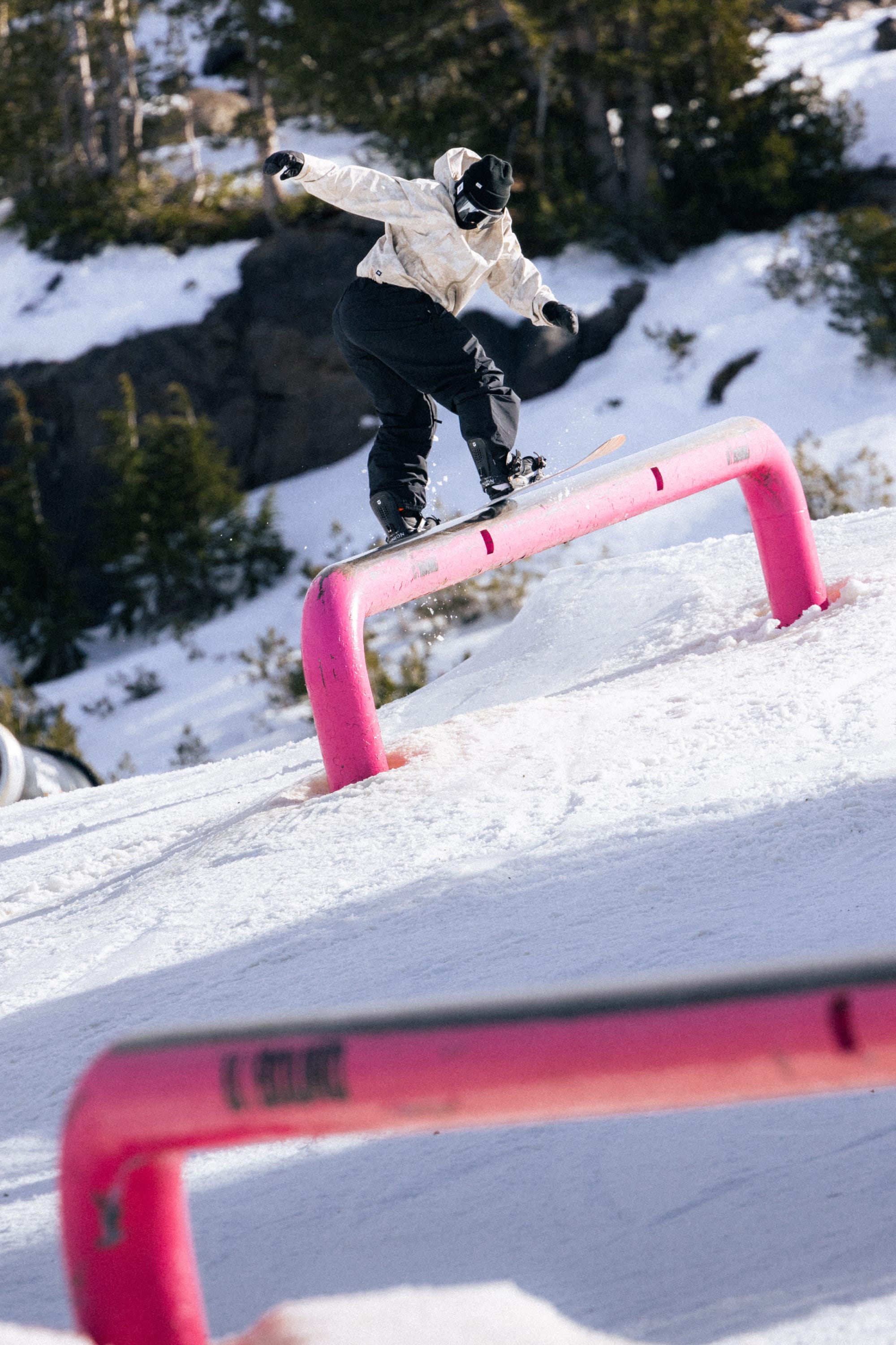 Wearing the 686 Women’s SMARTY® 3-in-1 Framework™ 3L Jacket, a snowboarder in black pants slides on a bright pink rail in a snowy terrain park surrounded by snow and trees.