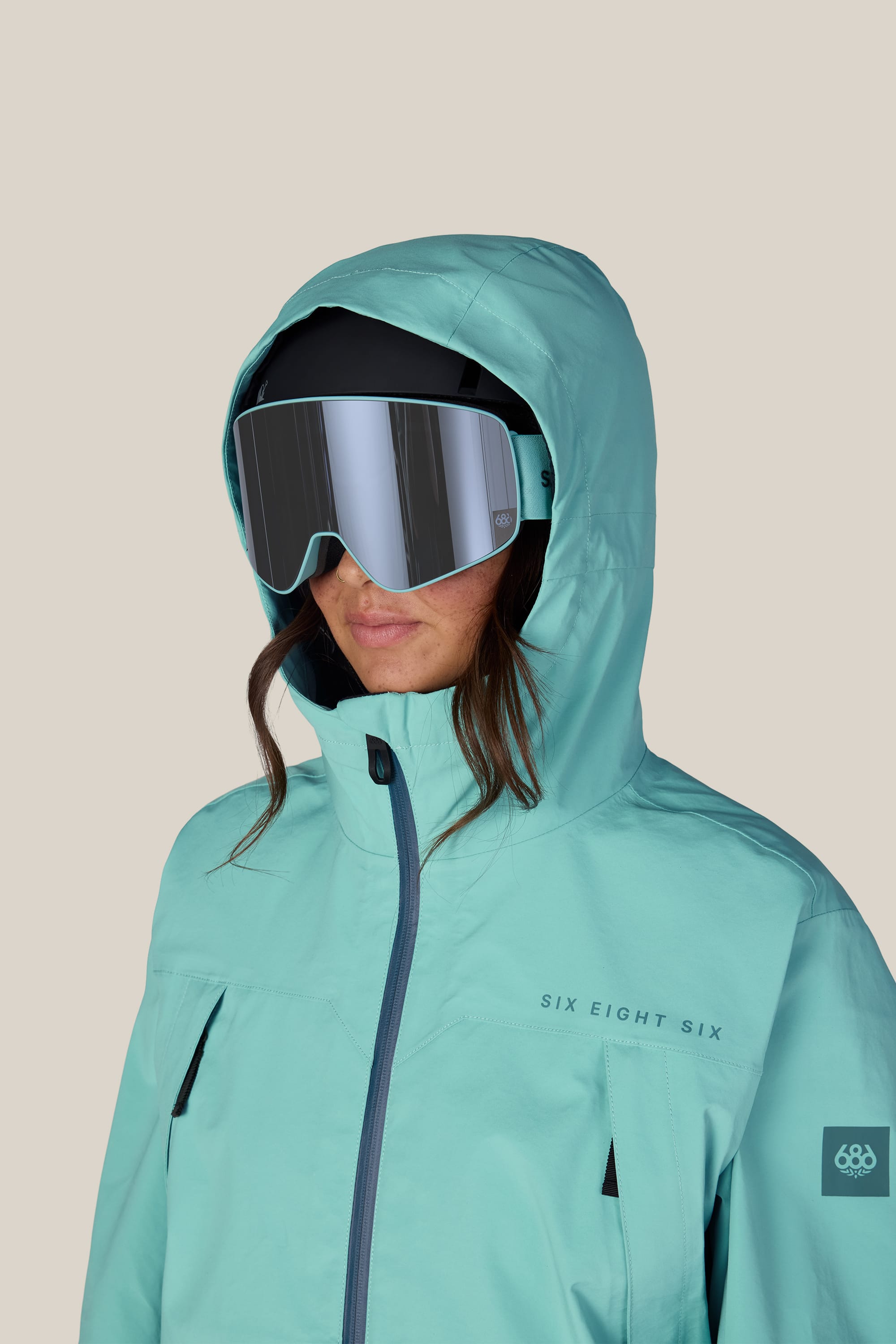 A person facing forward wears the 686 Women’s SMARTY® 3-in-1 Framework™ 3L Jacket in teal, featuring a hood and large reflective ski goggles. The waterproof polyester jacket displays “Six Eight Six” on the chest. The background is plain beige.