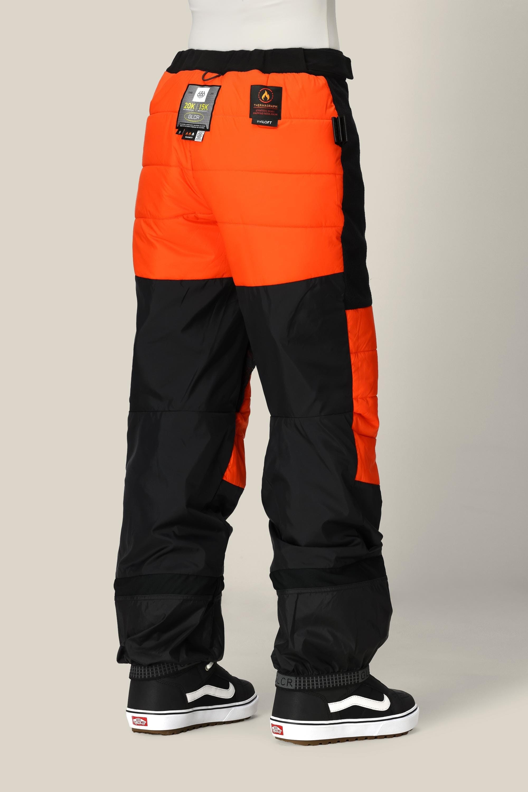 A person wearing black and orange 686 Women’s Geode Thermagraph® Pant with brand tags on the back, paired with black and white sneakers, standing against a neutral background.