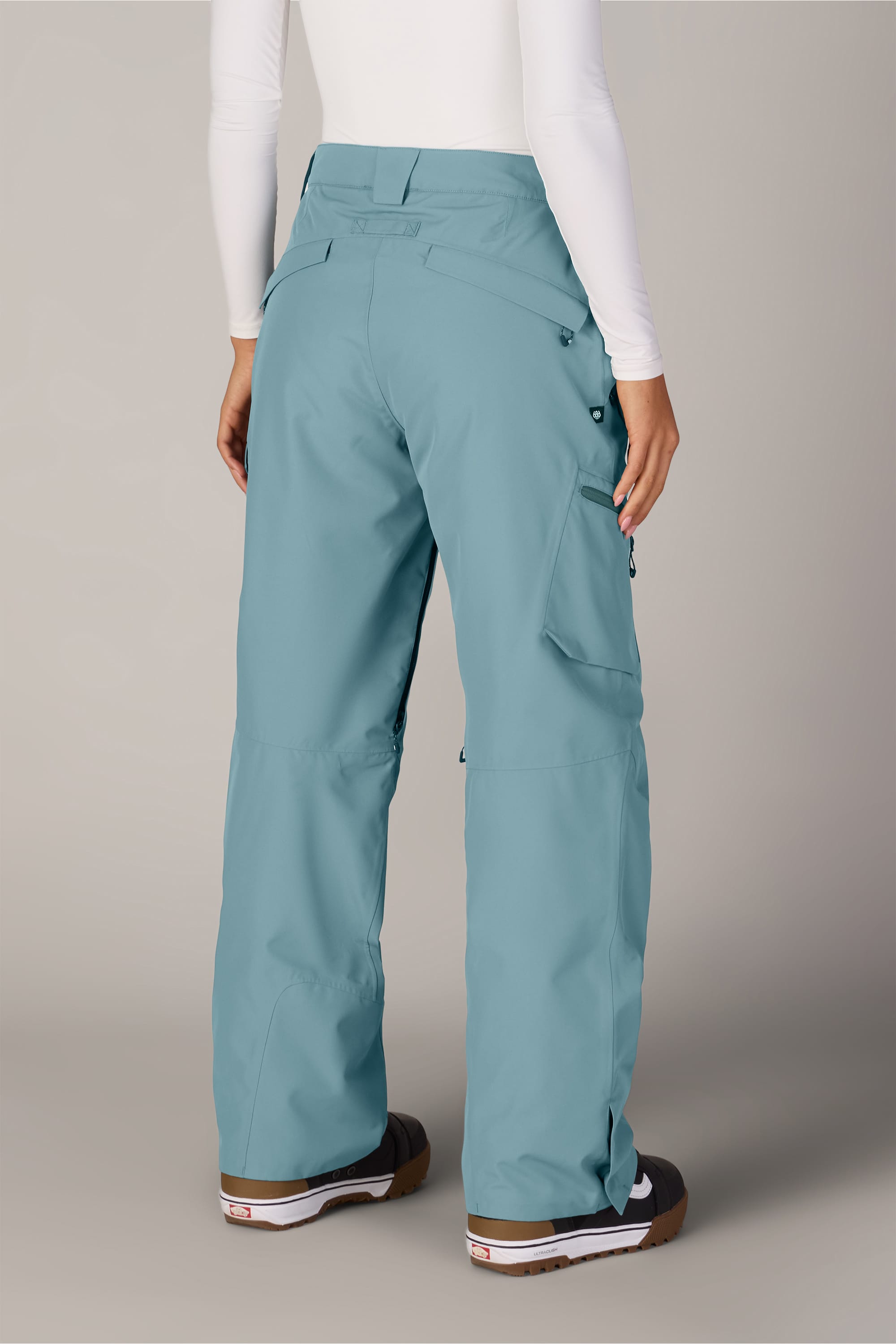 A person is seen from behind against a neutral background, wearing the 686 Women’s Geode Thermagraph® Pant in light blue by 686, paired with brown boots and a white long-sleeve top.