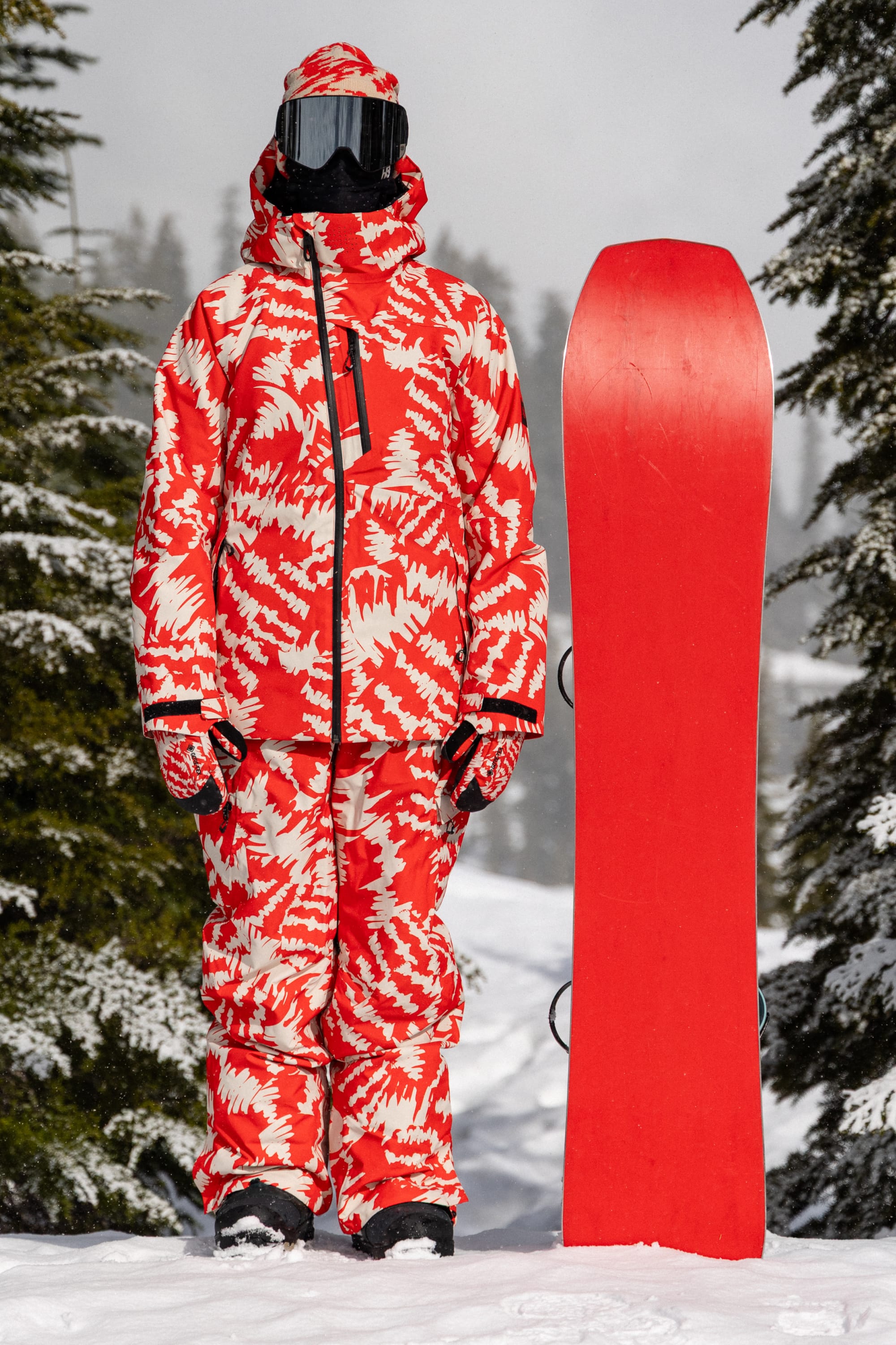 A person in a red and white patterned snowsuit and black goggles stands on snow next to a plain red snowboard, with pine trees and snow in the background, their outfit complemented by the 686 Women’s Geode Thermagraph® Pant from 686, designed for maximum comfort and protection on the slopes.