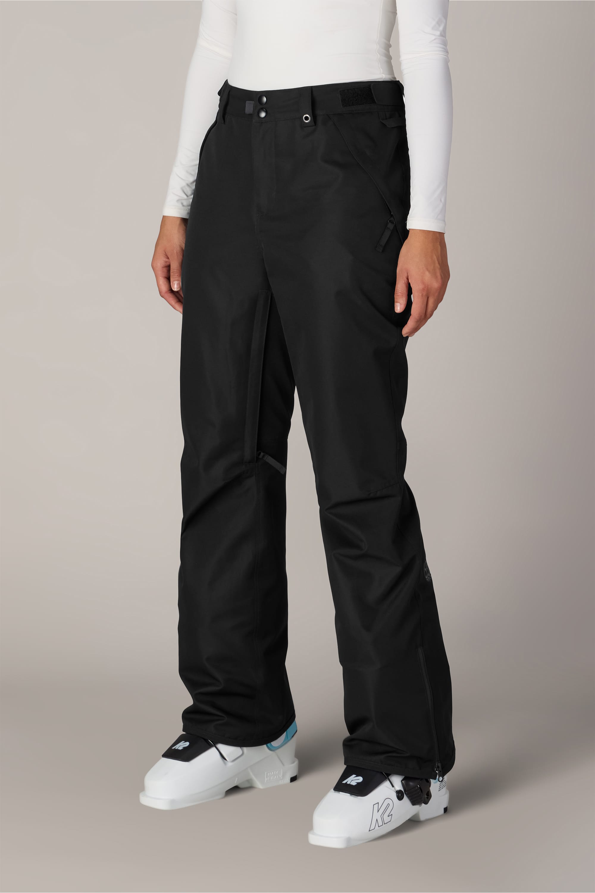 A person is wearing the 686 Women’s Slim Line Pant in black polyester, paired with a white long-sleeve top and white ski boots, standing against a plain light background.