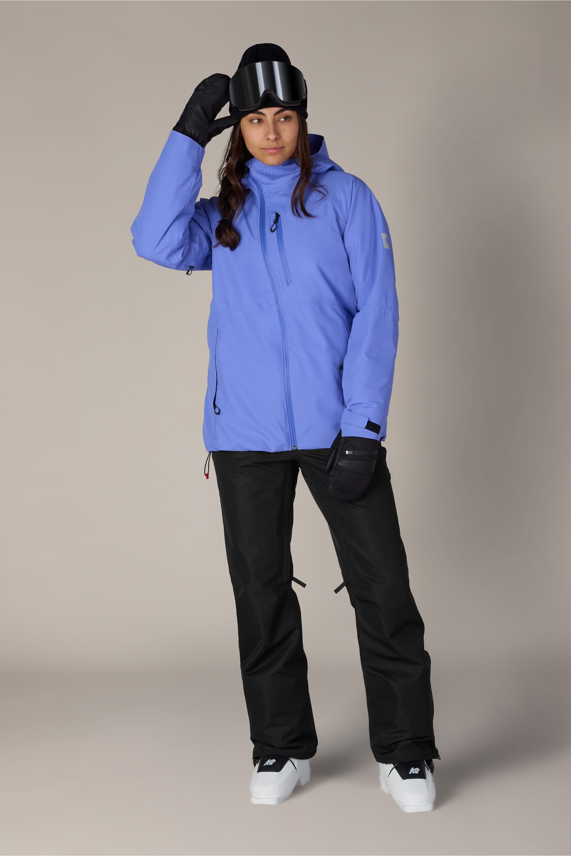 A person wearing the 686 Women’s Slim Line Pant by 686, paired with a light blue jacket, gloves, and white boots stands holding a black ski helmet and goggles, looking at the camera.