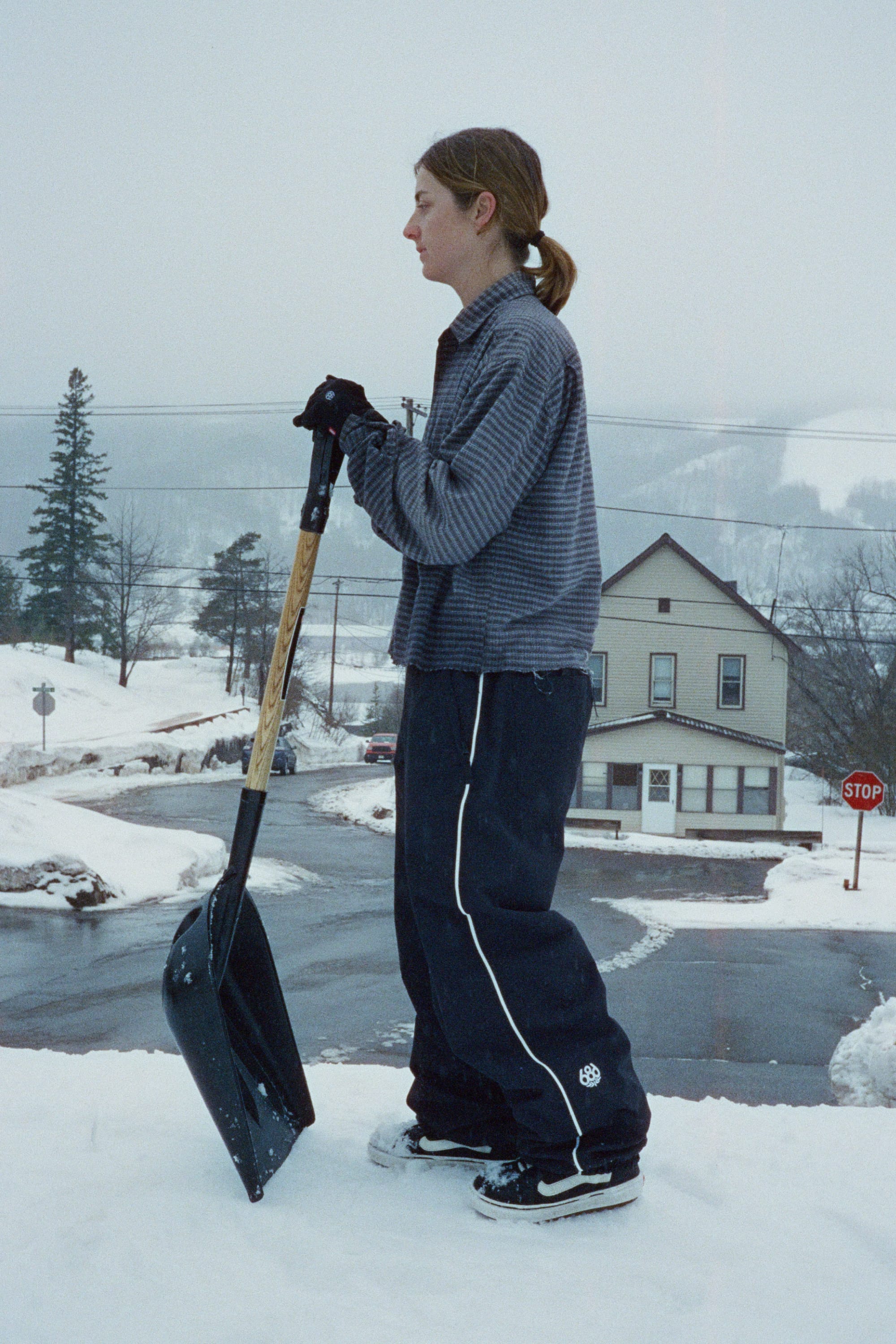 A person in winter clothes stands on snow holding a large shovel, dressed in the 686 Women’s Hellkat™ Shell Pant by 686, with snowy streets, houses, and trees in the background on a cloudy day.