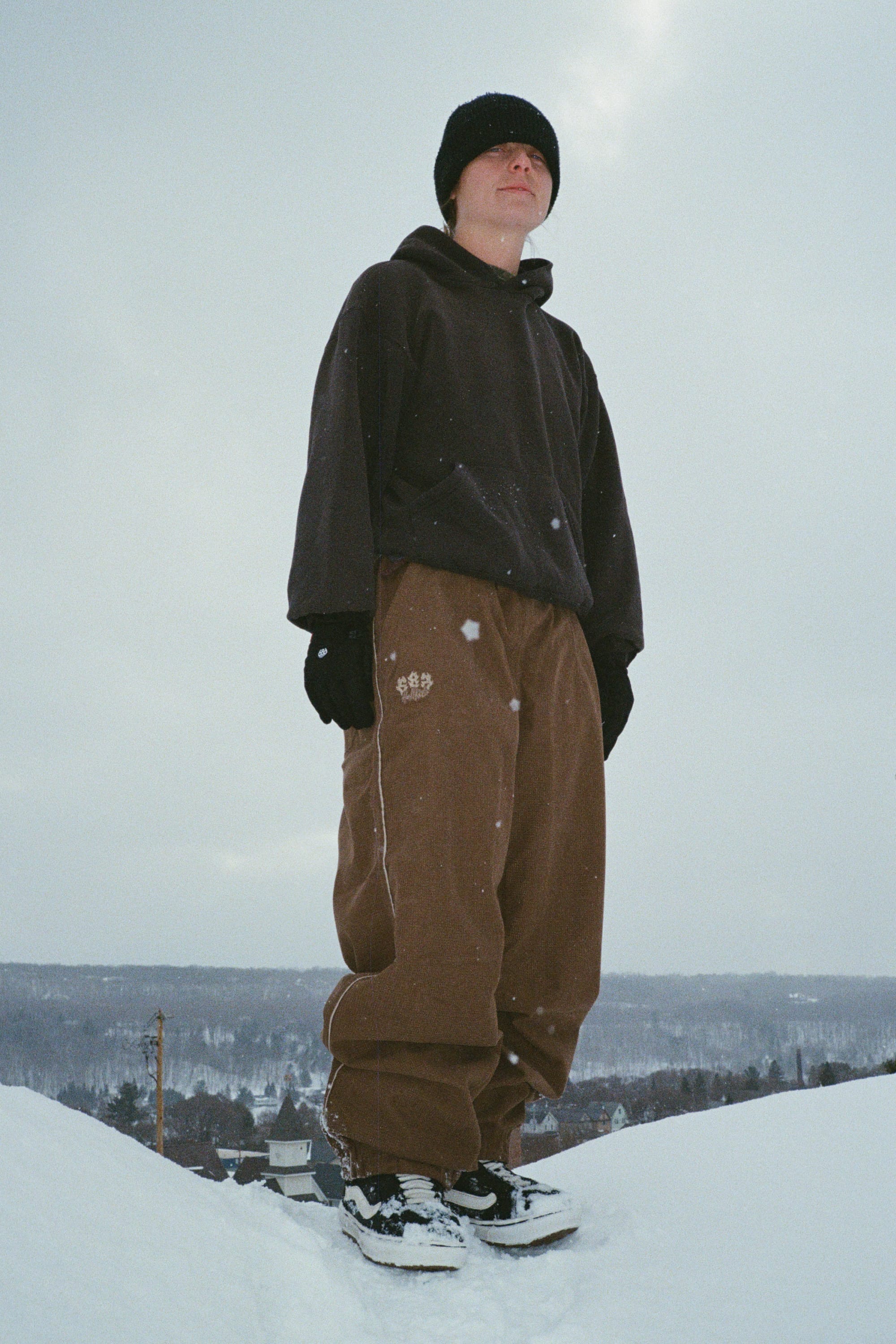 A person in a black beanie, black hoodie, 686 Women’s Hellkat™ Shell Pant by 686, and gloves stands on a snowy hill with a cloudy sky and snowy landscape in the background.