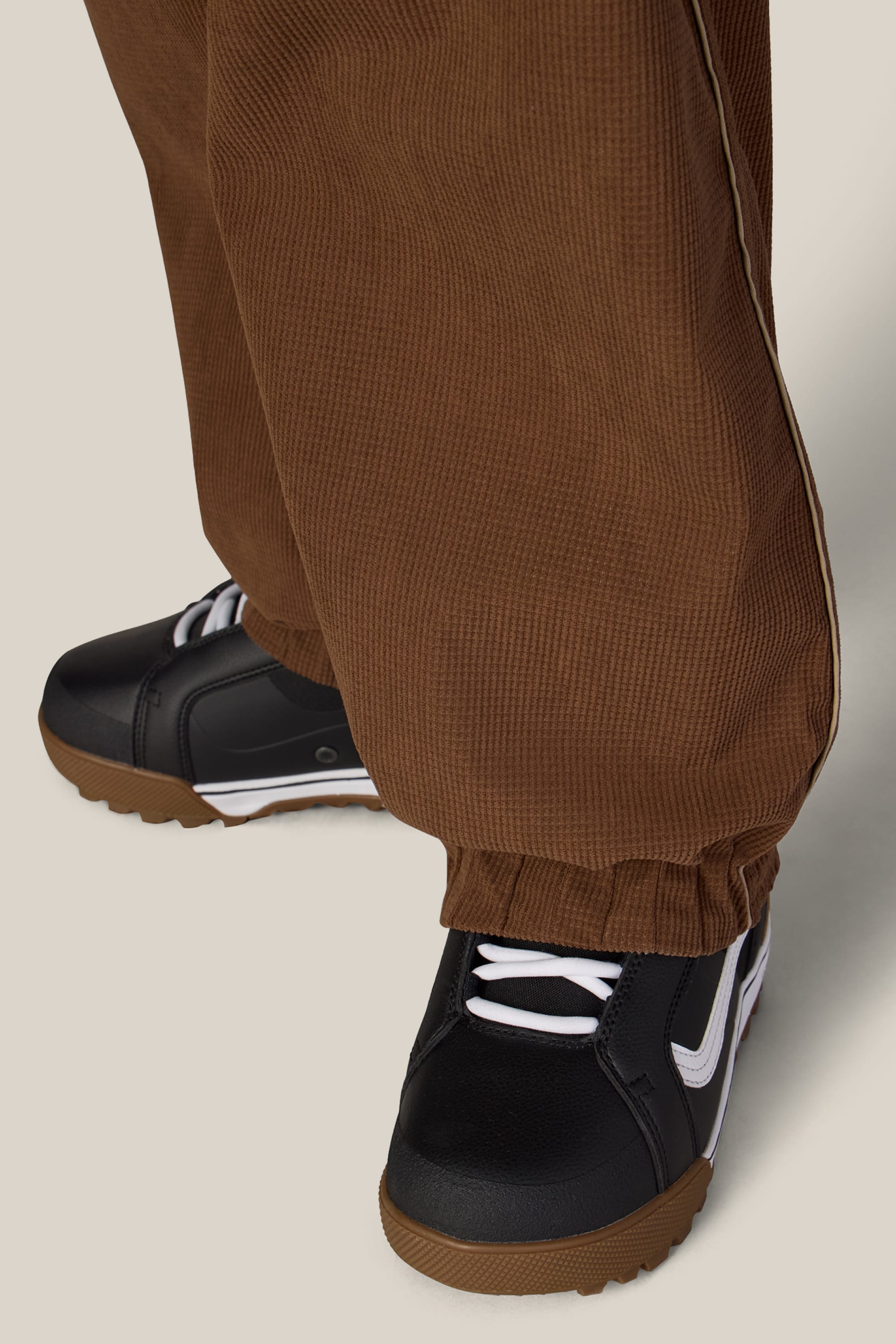 A person is wearing the 686 Women’s Hellkat™ Shell Pant in brown, paired with black sneakers featuring white details, standing on a light beige surface. The pants from 686, styled like women’s woven pants, partially cover the sneakers.