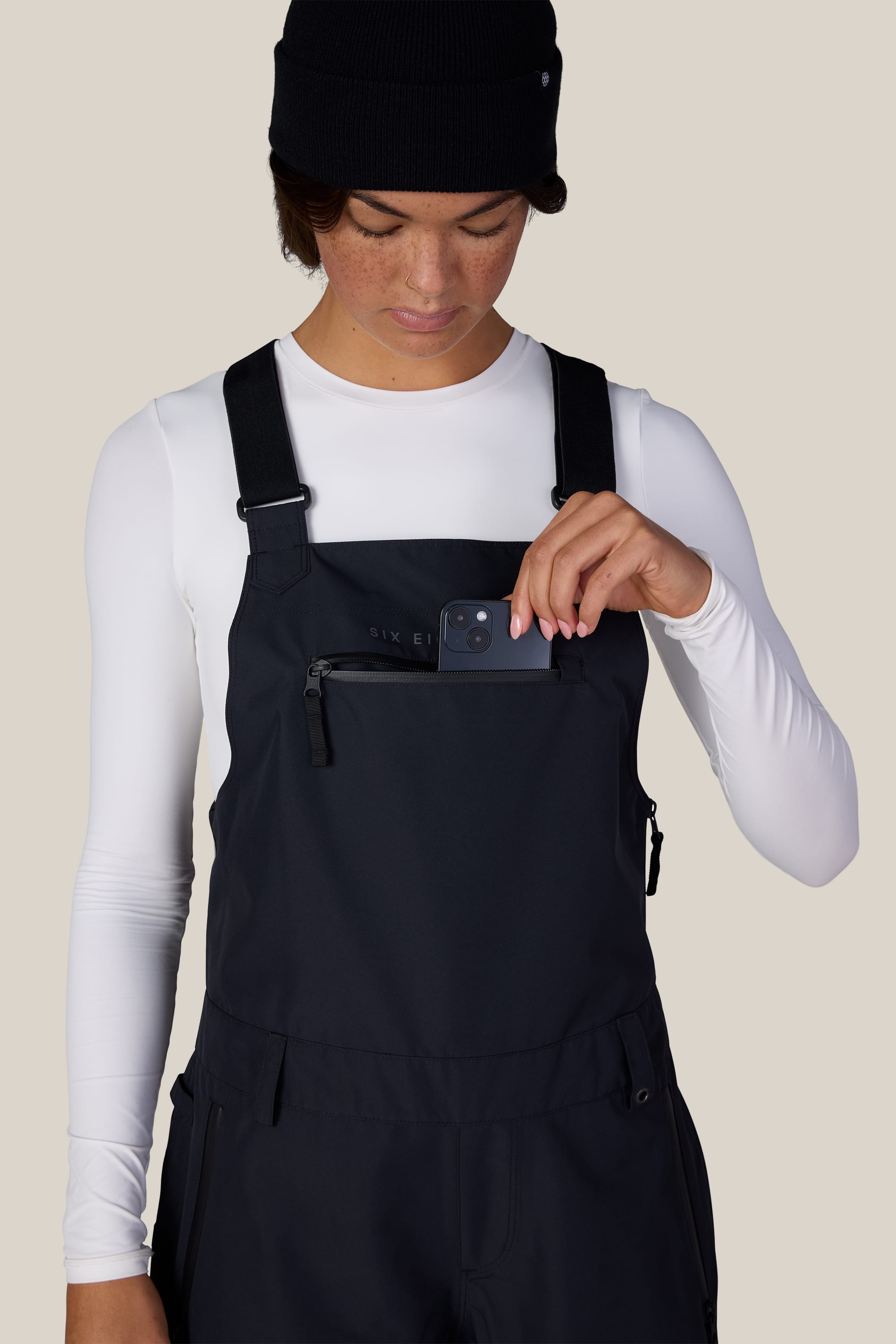 A person wearing a white long-sleeve shirt, the 686 Women’s Regalia™ Bib by 686 in black, and a black beanie places a small black device into the zippered chest pocket of the overalls, against a light beige background.