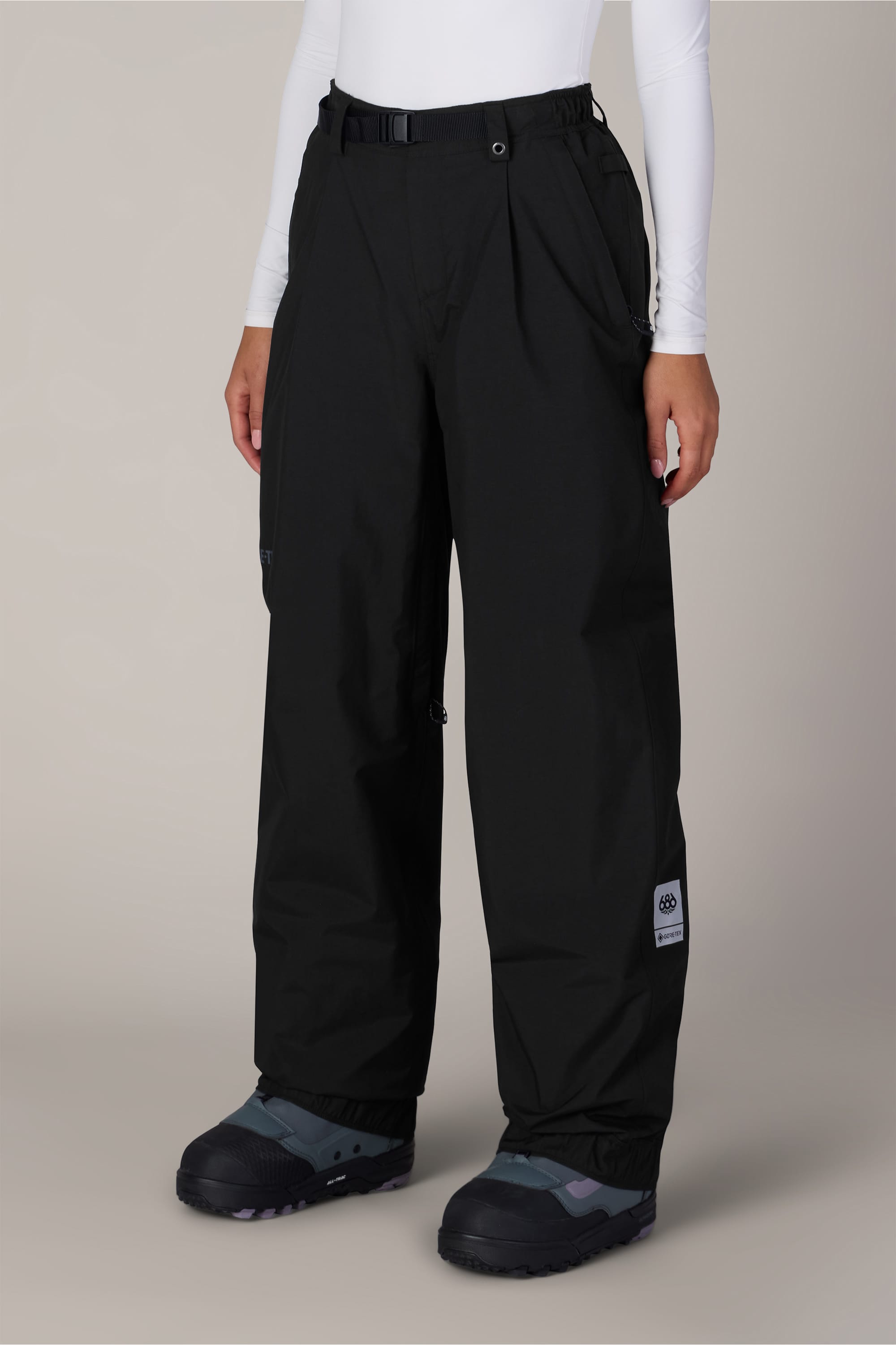 A person wearing 686 Women’s GORE-TEX® Outline™ Pant in black, paired with black winter boots and a white long-sleeve top, stands against a plain light background. Only the lower half of the body is visible.