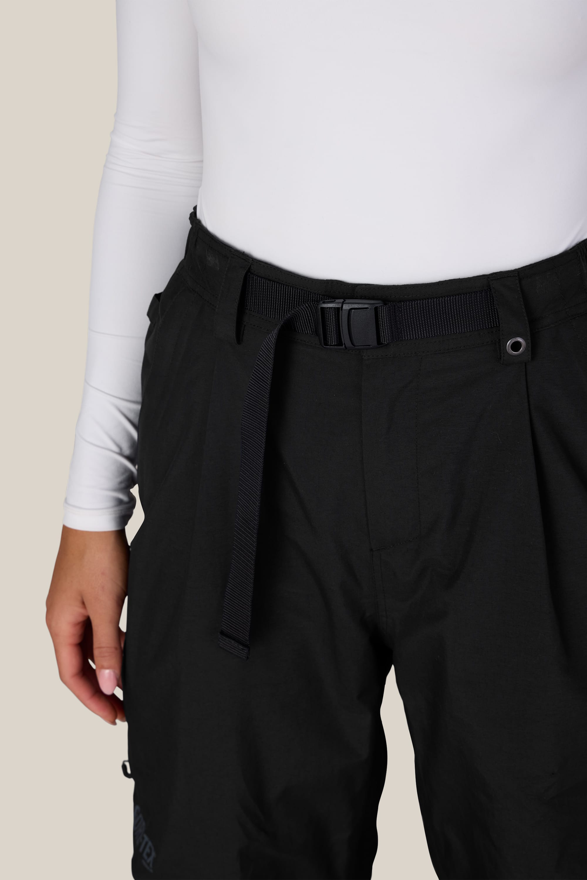 A person wearing a white long-sleeve top and the 686 Women’s GORE-TEX® Outline™ Pant by 686, featuring a black high-waisted design with a black belt, stands against a plain background. Only the torso and upper legs are visible.