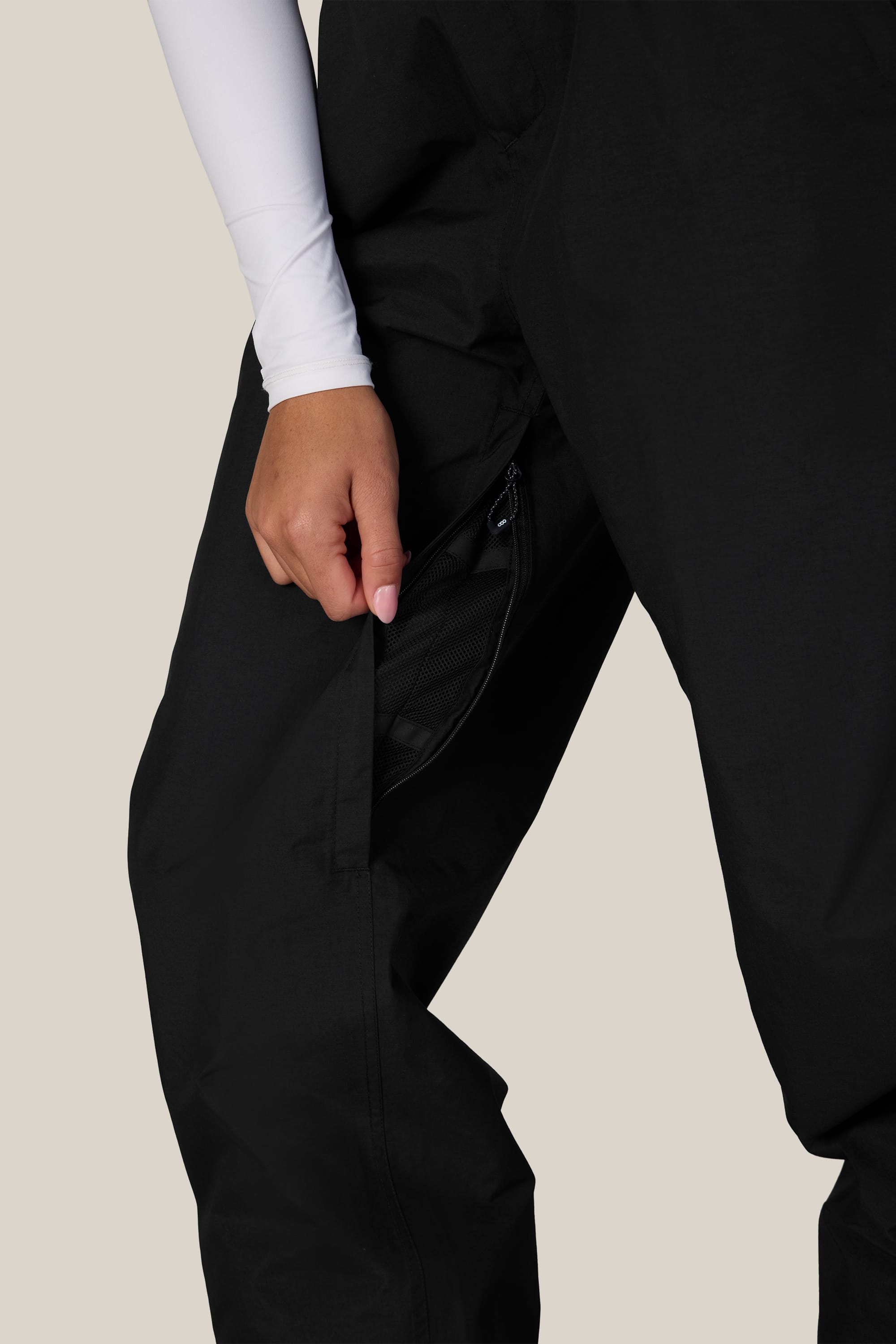 A person wearing the 686 Women’s GORE-TEX® Outline™ Pant in black and a white long-sleeve shirt opens a zippered pocket on the side of their pants.