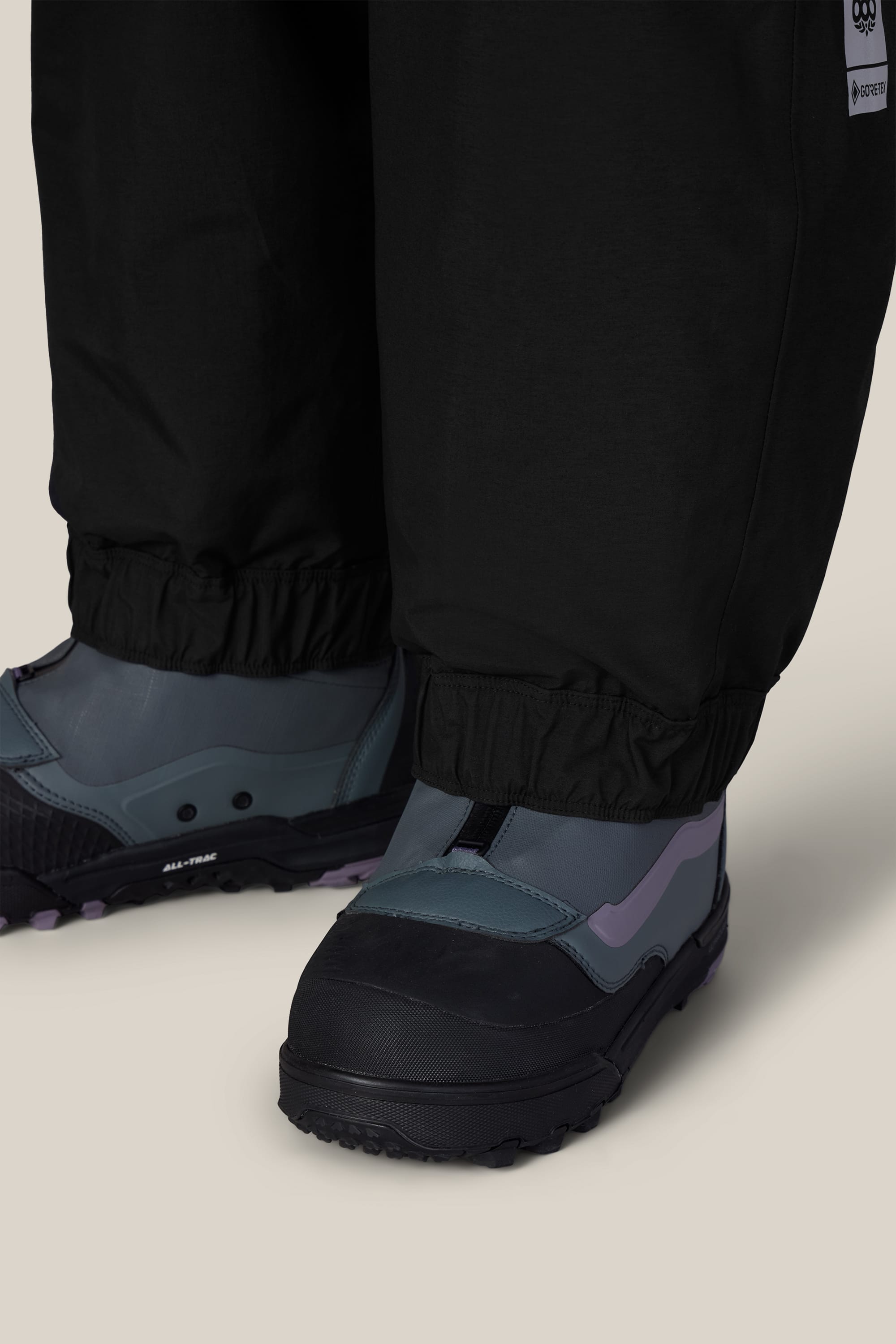 Close-up of a person wearing the 686 Women’s GORE-TEX® Outline™ Pant in black with elastic cuffs, paired with gray-blue winter boots, on a light background.