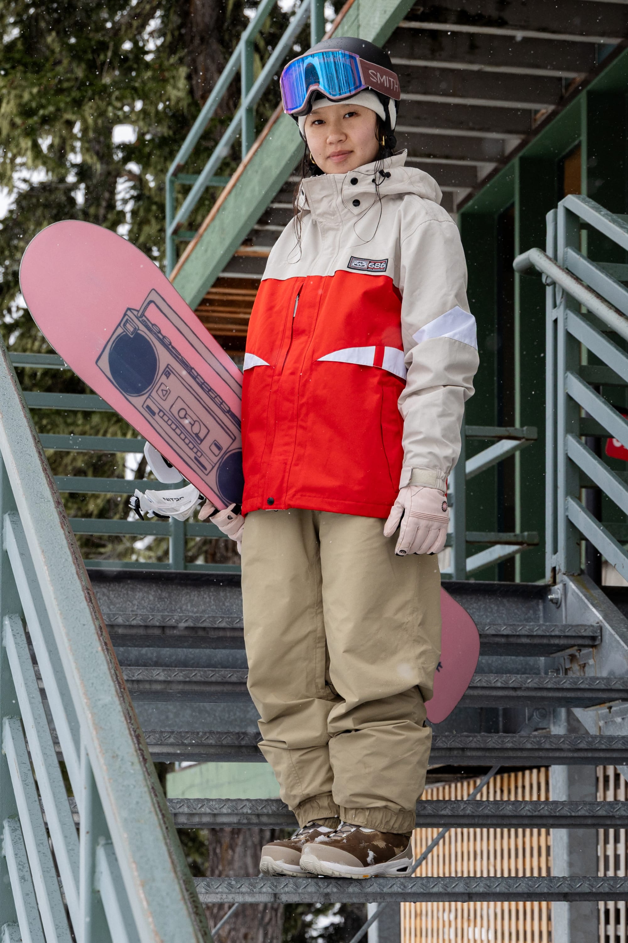A snowboarder in a red and beige jacket, 686 Women’s GORE-TEX® Outline™ Pant, and goggles stands on outdoor stairs holding a pink snowboard with a boombox graphic. Snow and trees are in the background.