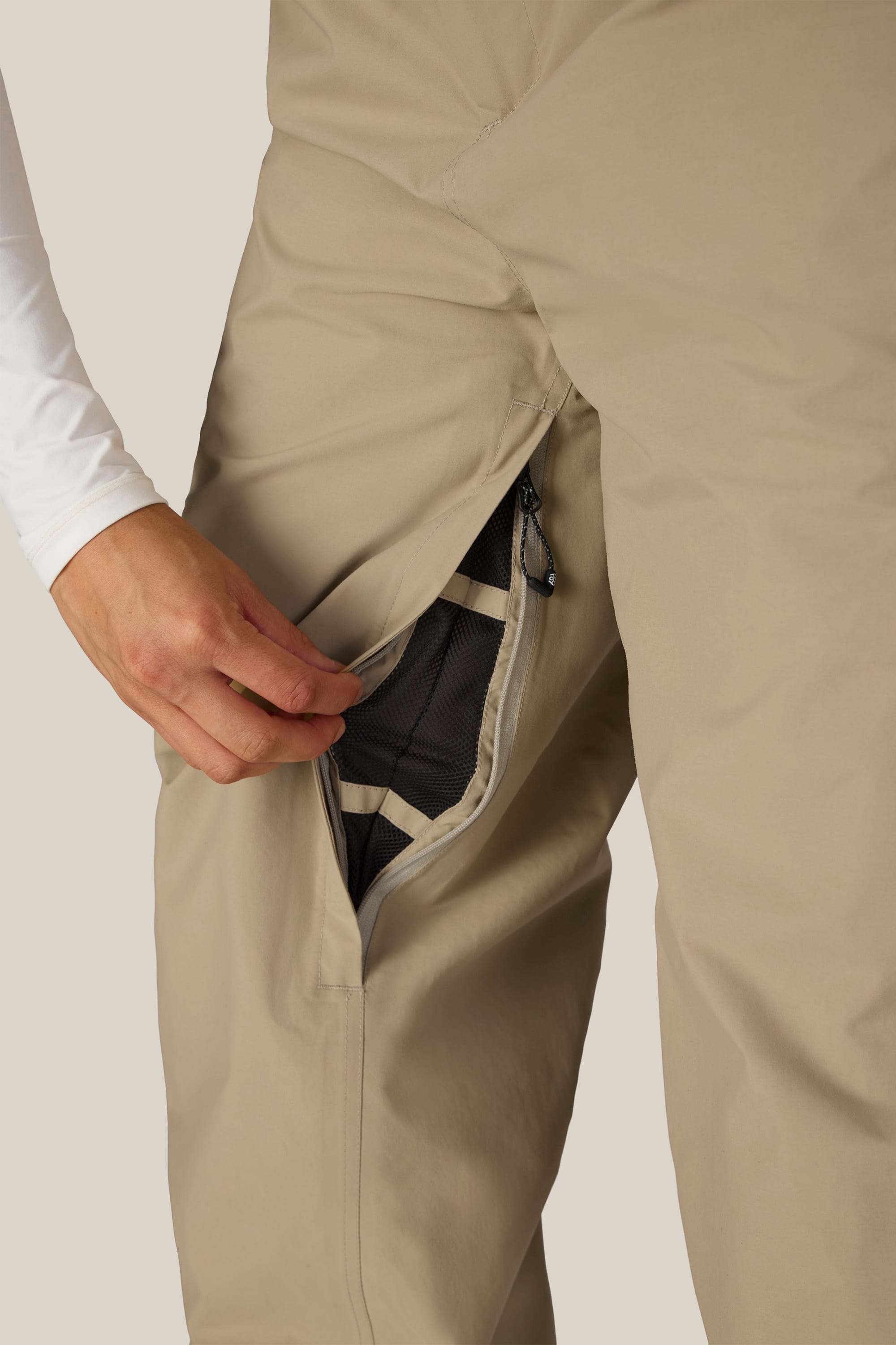 A woman wearing 686 Women’s GORE-TEX® Outline™ Pants in beige unzips the thigh vent, revealing the mesh lining inside. She pairs the pants with a long-sleeve white shirt.